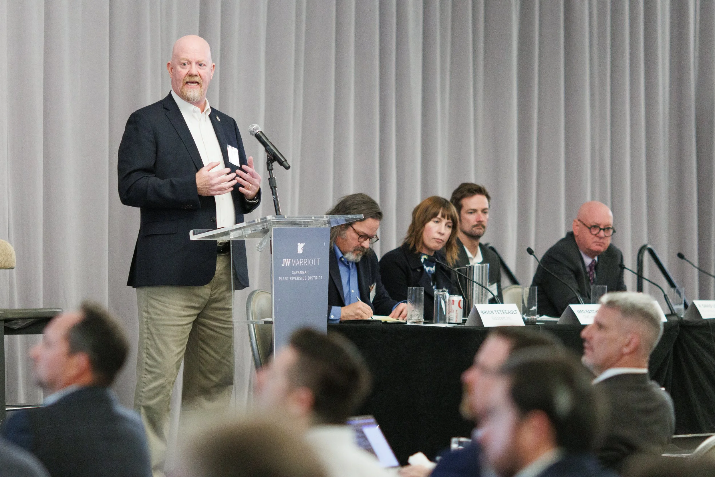 A man in a black blazer and white shirt speaking at a podium with a microphone at a conference. Four seated panelists, two men and two women, are listening. Audience members are visible in the foreground.