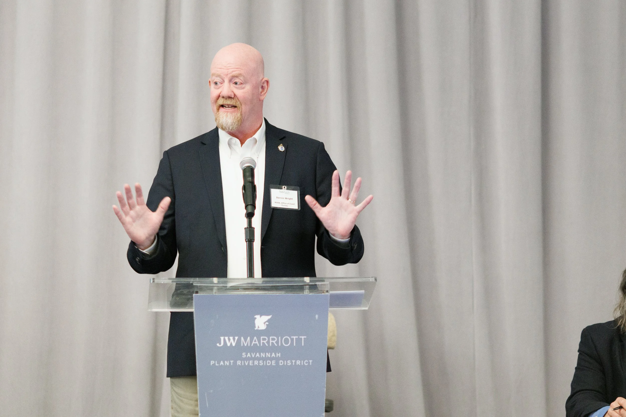 A man with a beard and bald head wearing a dark blazer and white shirt standing behind a podium with a JW Marriott sign. He is speaking into a microphone and gesturing with his hands against a gray curtain background.