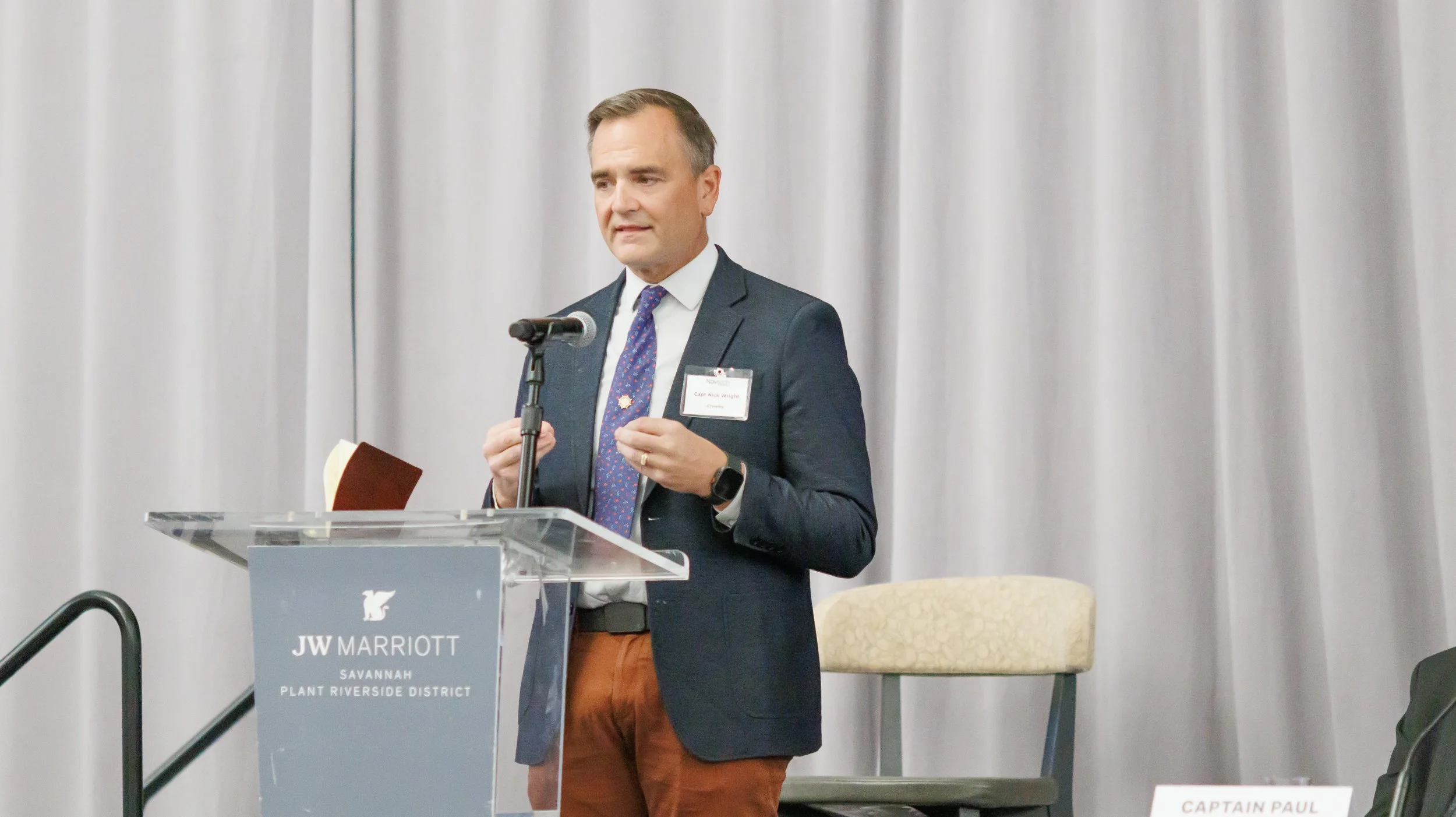 Man in a suit speaking at a podium with a microphone at a conference. He is wearing a name badge and looking slightly to the side. The podium has a sign that reads 'JW Marriott Savannah Plant Riverside District'.
