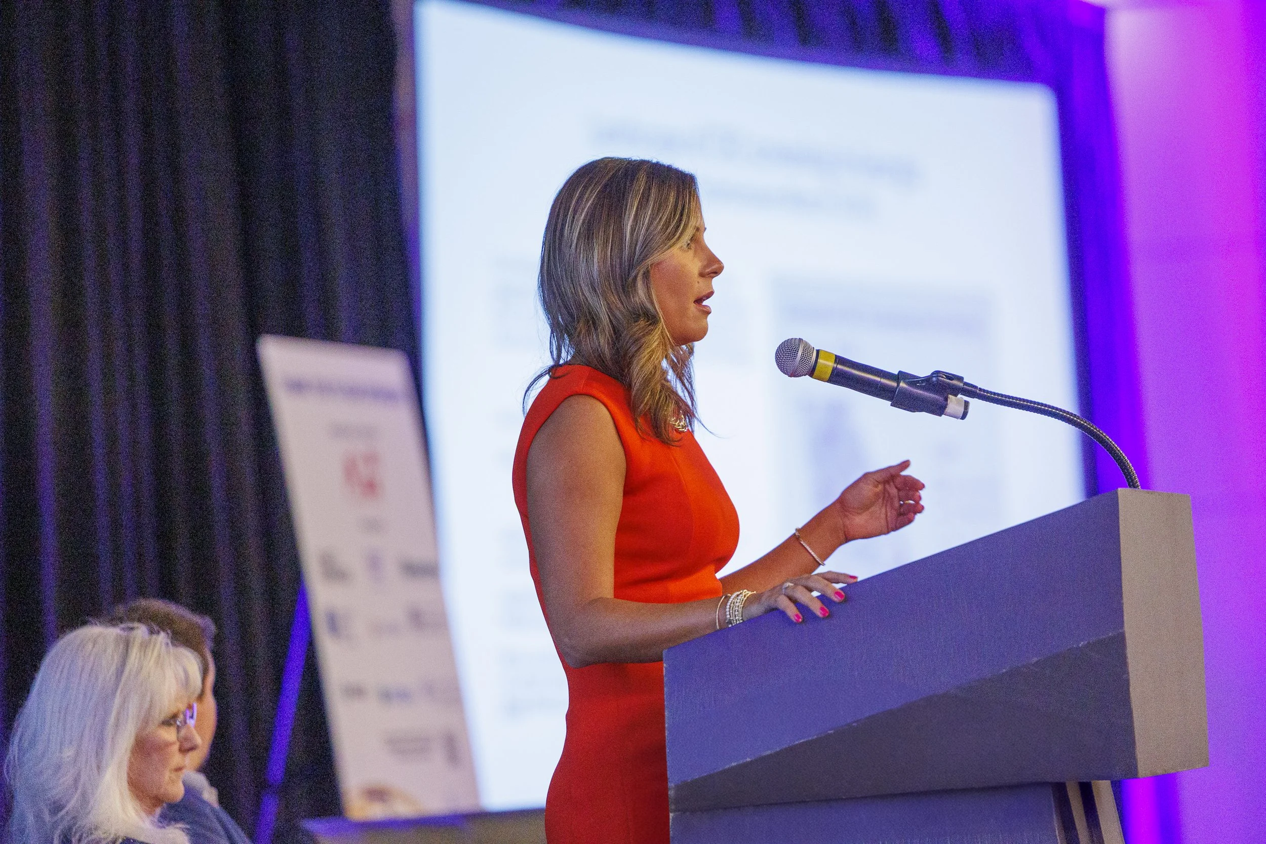 A woman in a red dress speaking at a podium during a conference, with a microphone and a presentation screen behind her.