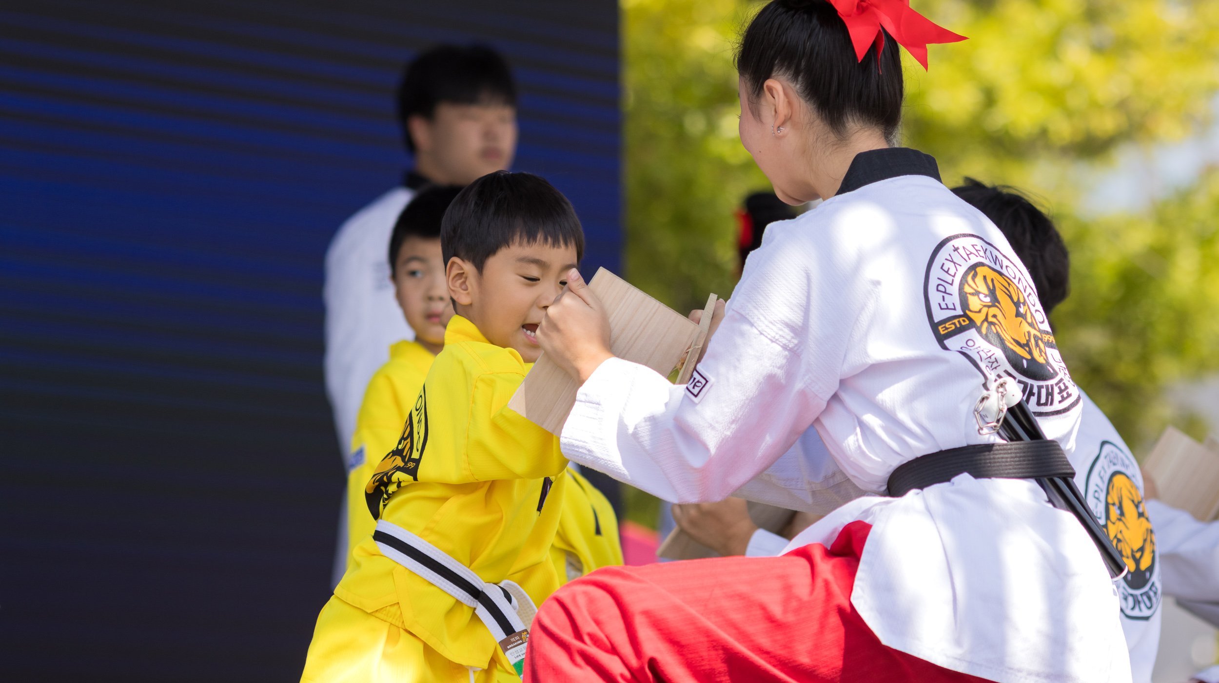 A girl in a white martial arts uniform with a black belt and red bow giving a medal to a boy in a yellow jacket during an outdoor martial arts event, with other children in the background.