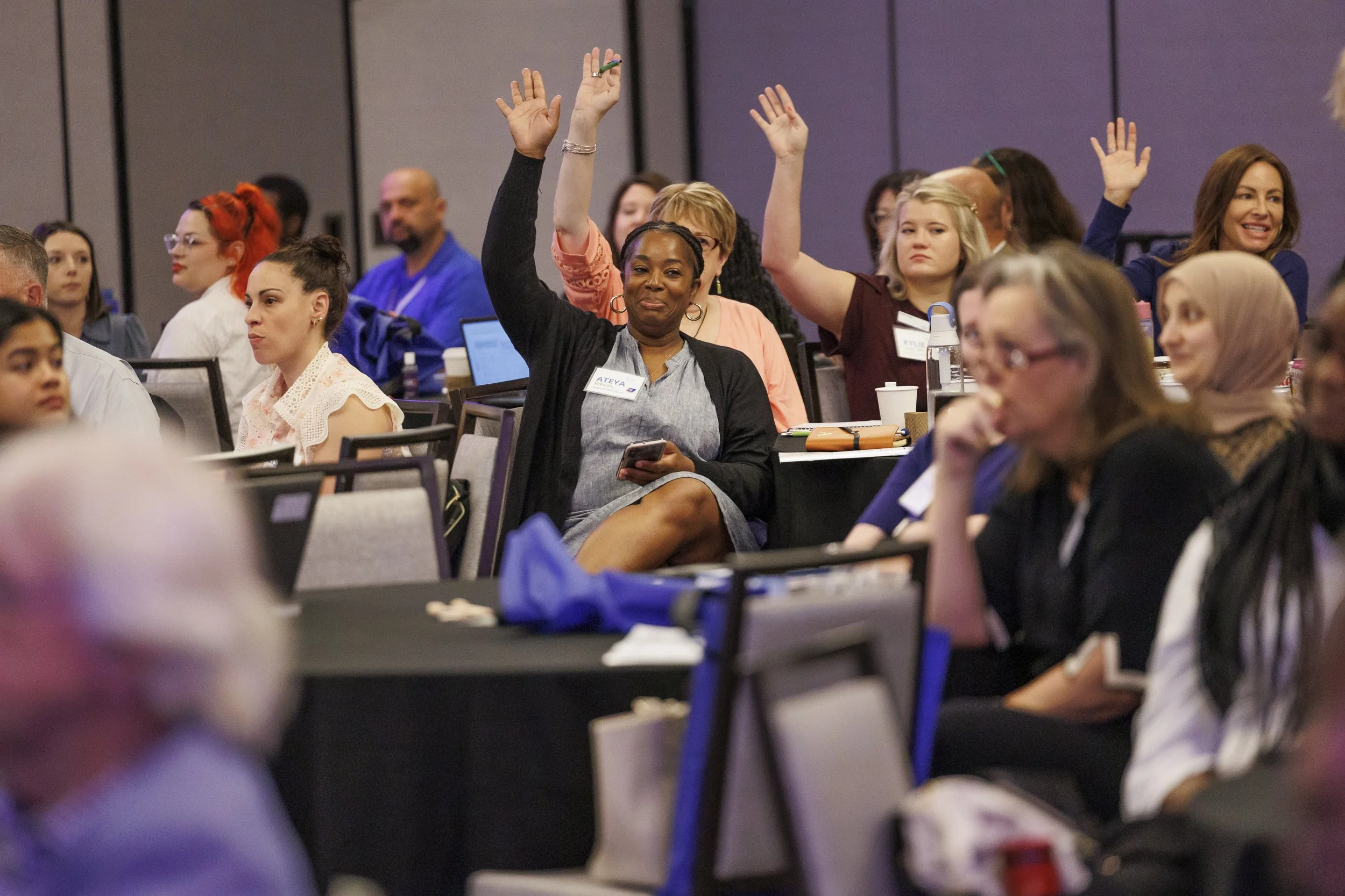A diverse group of people at a conference, some raising their hands, seated at tables with laptops and notepads.