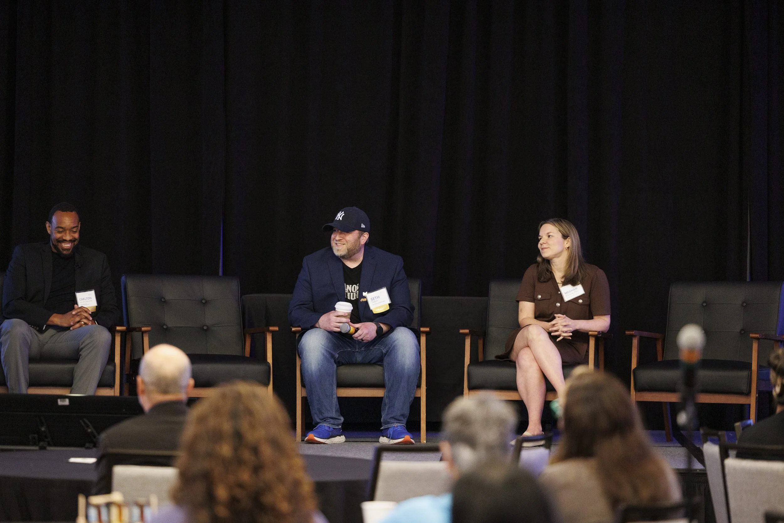 Panel discussion with three people on stage, sitting on black chairs, with an audience in front. The man in the middle is holding a coffee cup and wearing a navy blazer and New York Yankees cap. The woman on the right is in a brown dress with crossed