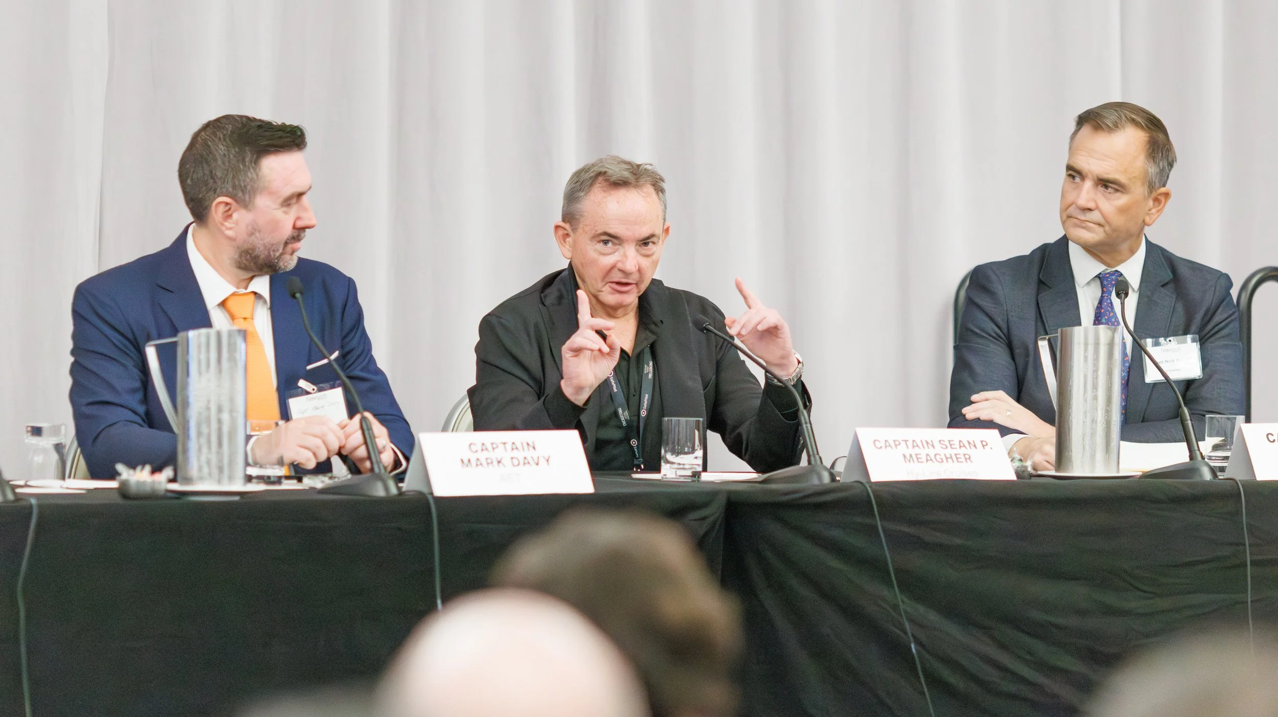 Three men sitting at a panel discussion table with microphones. The man in the middle is speaking, with nameplate indicating he is Captain Sean P. Meagher. The man on the left is wearing a dark blue suit and orange tie, and the man on the right is we