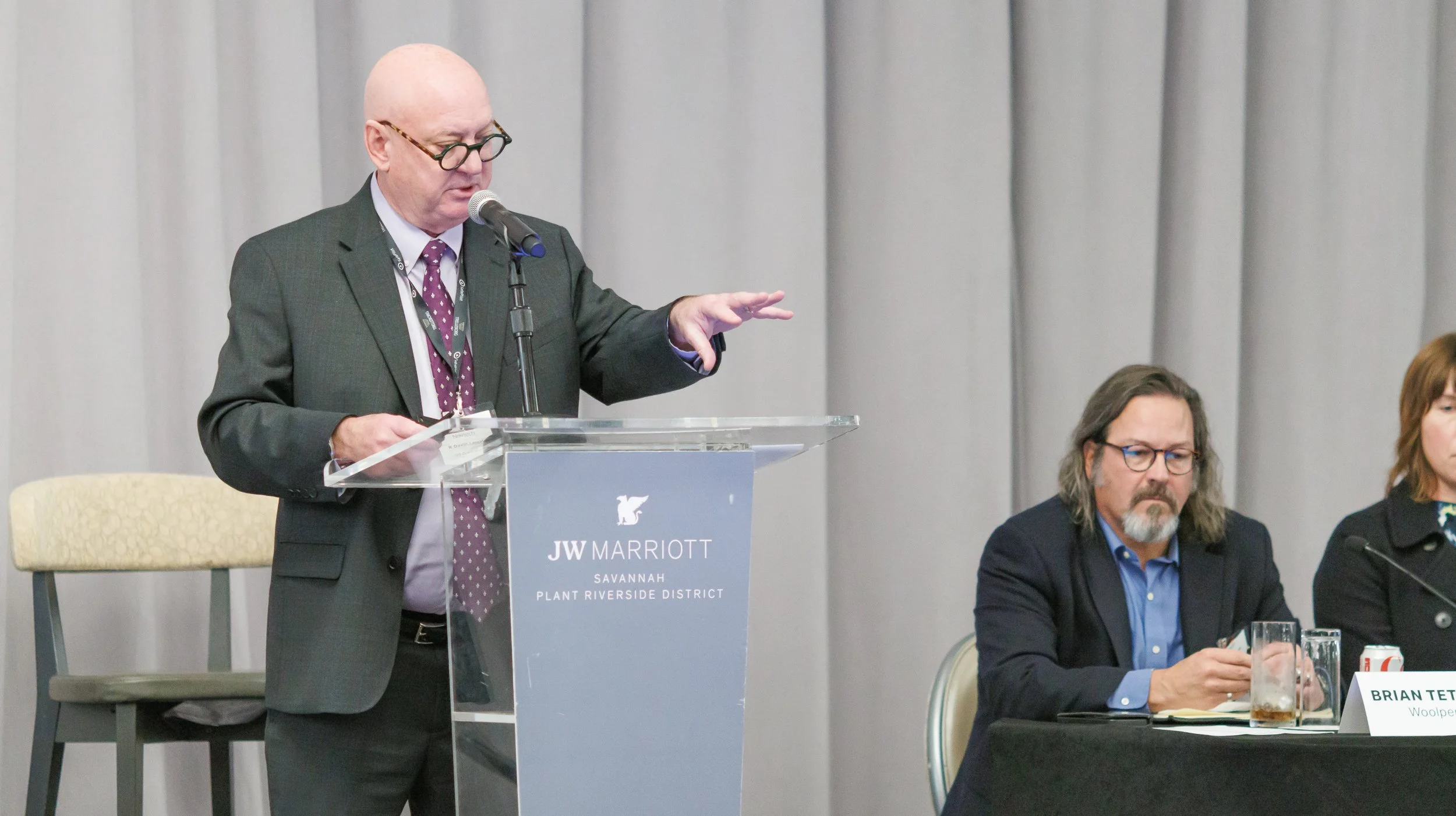 A man in a suit and glasses speaking at a podium with a microphone at the JW Marriott Savannah Plant Riverside District. Two seated individuals, a man with long hair, glasses, a beard, and a woman, are listening and have drinks on the table.