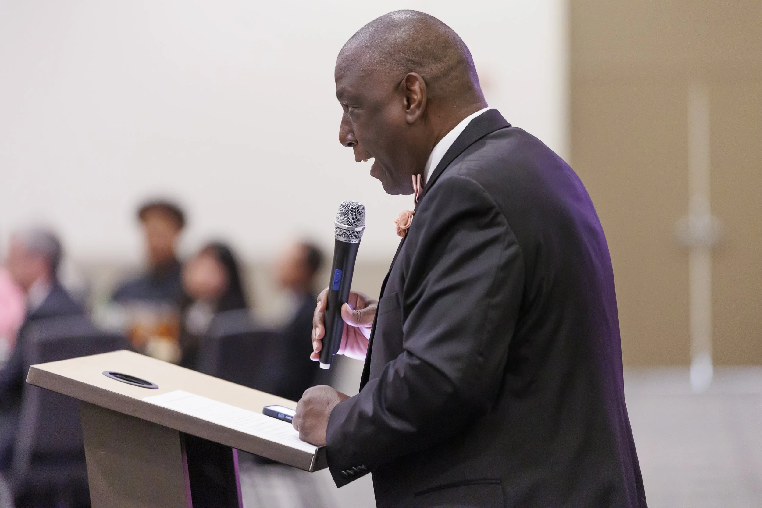 A man in a black tuxedo giving a speech at a podium while holding a microphone at a formal event.