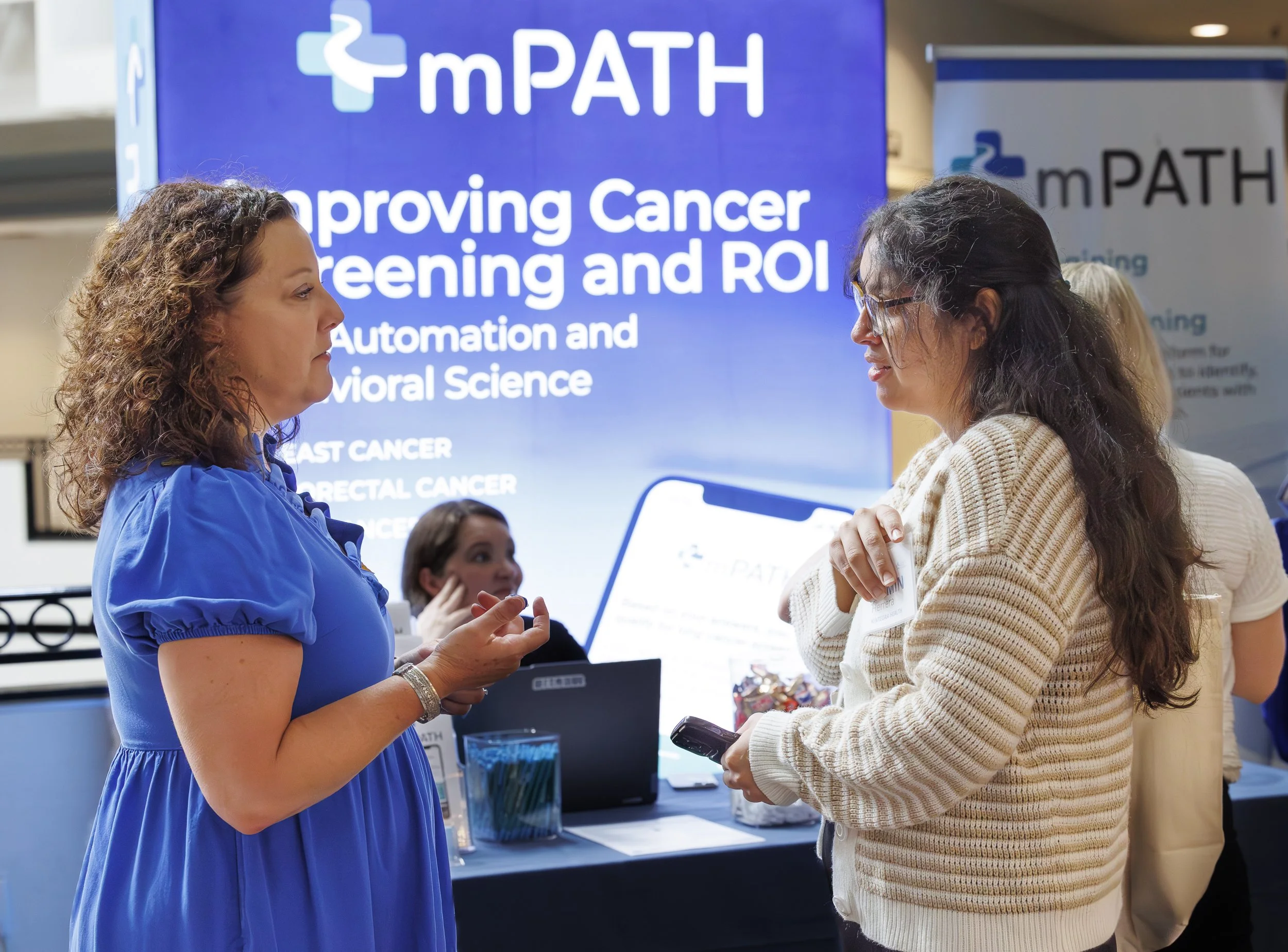 Two women conversing at a medical conference booth with a large blue sign behind them that reads 'mPATH' and 'Improving Cancer Screening and ROI'.