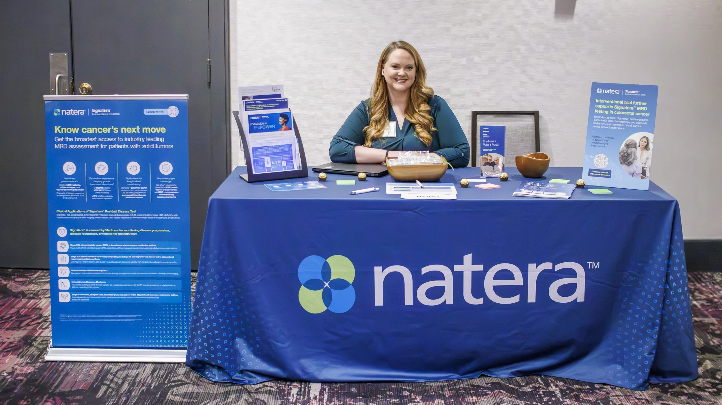 A woman seated at a Natera informational booth with a blue tablecloth featuring the Natera logo. The booth displays informational posters, pamphlets, and promotional items, with a large blue poster on a stand to her left and a smaller blue sign on th
