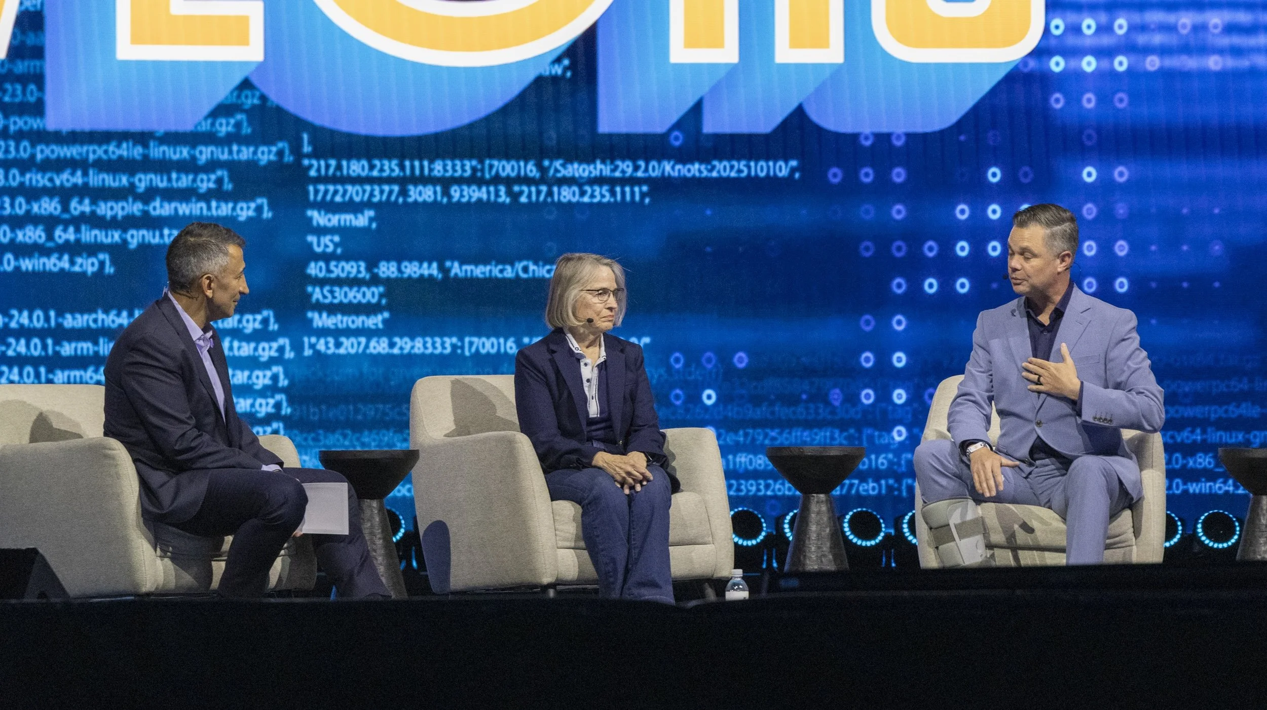 Three people seated on stage during a panel discussion with a large digital blue screen behind them displaying digital code.