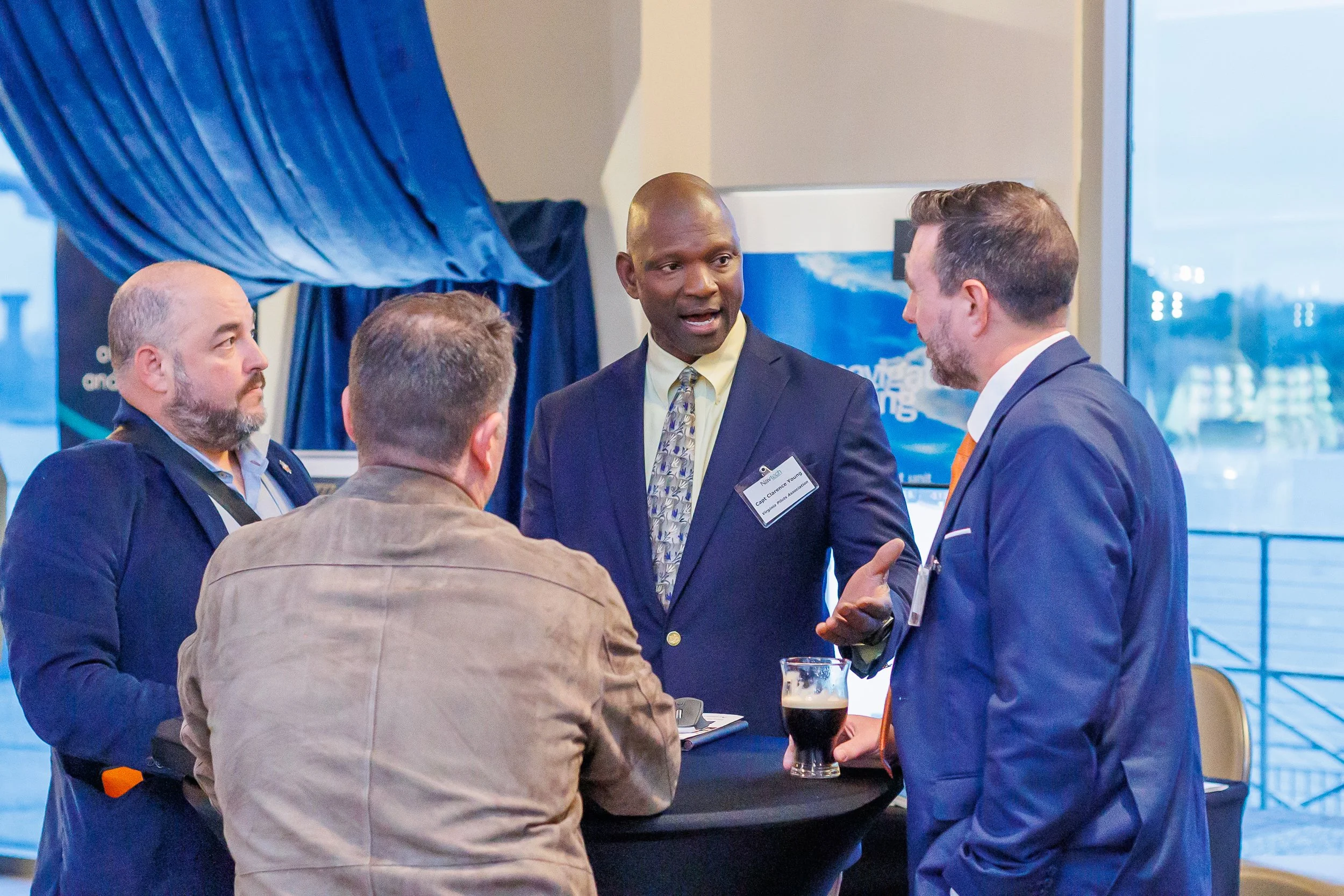 Four men in business attire are having a conversation at a networking event, with one man gesturing while talking and a glass of dark beer on the table.