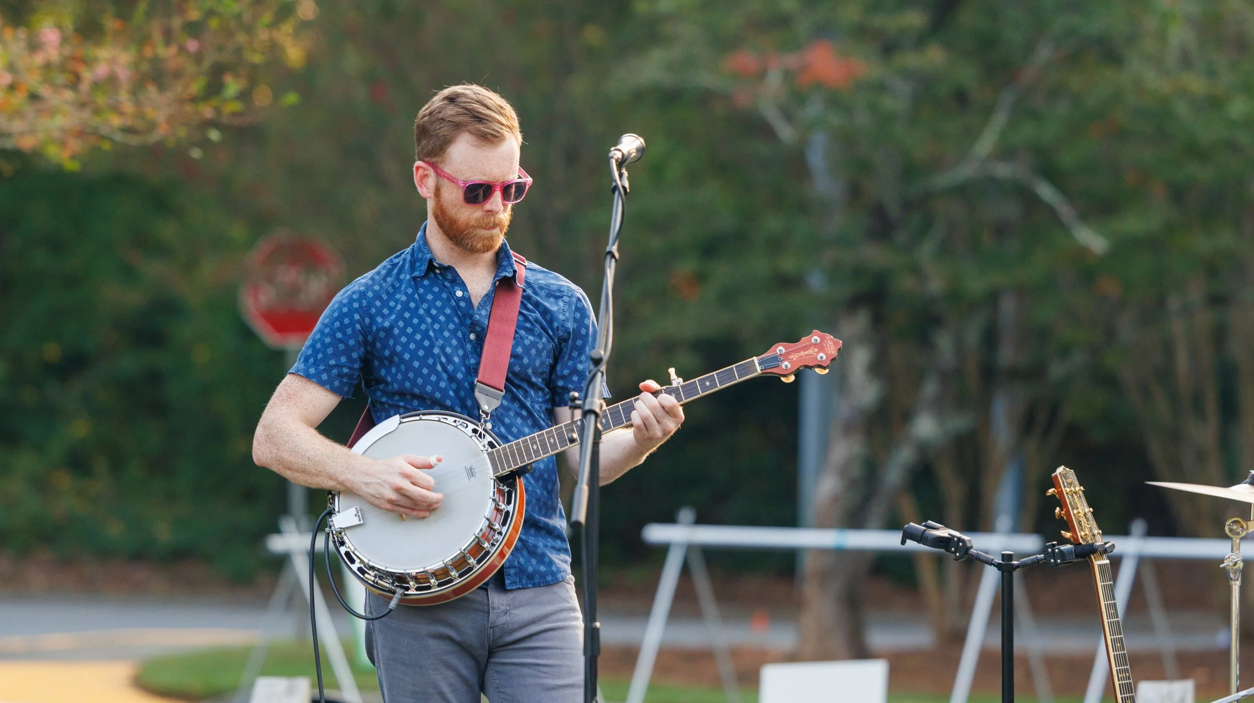 A man wearing pink sunglasses and a blue patterned shirt playing a banjo outdoors with trees in the background.