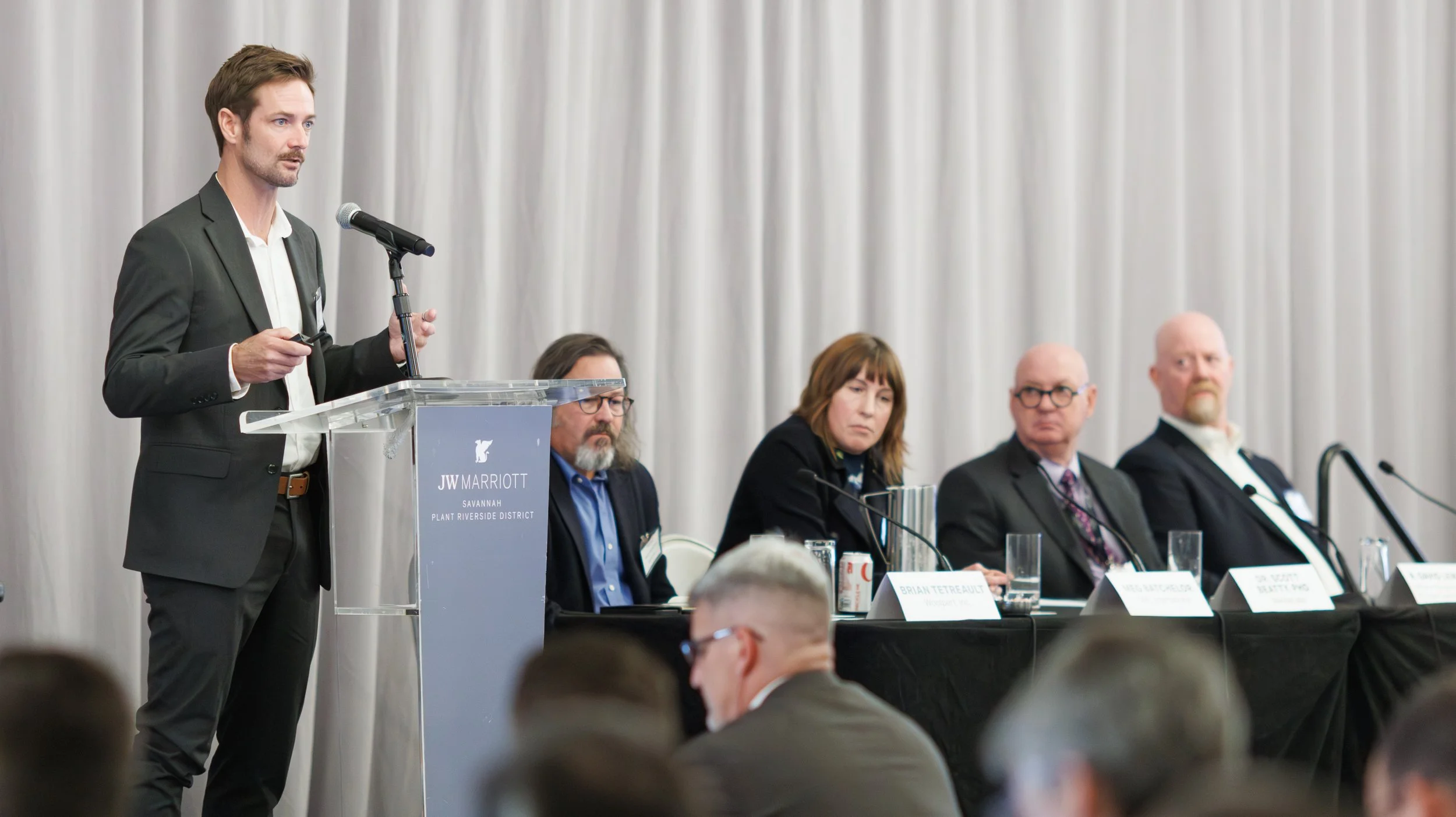 A man in a suit giving a presentation at a conference with a panel of four people seated behind him at a long table. The conference is at JW Marriott, Savannah.