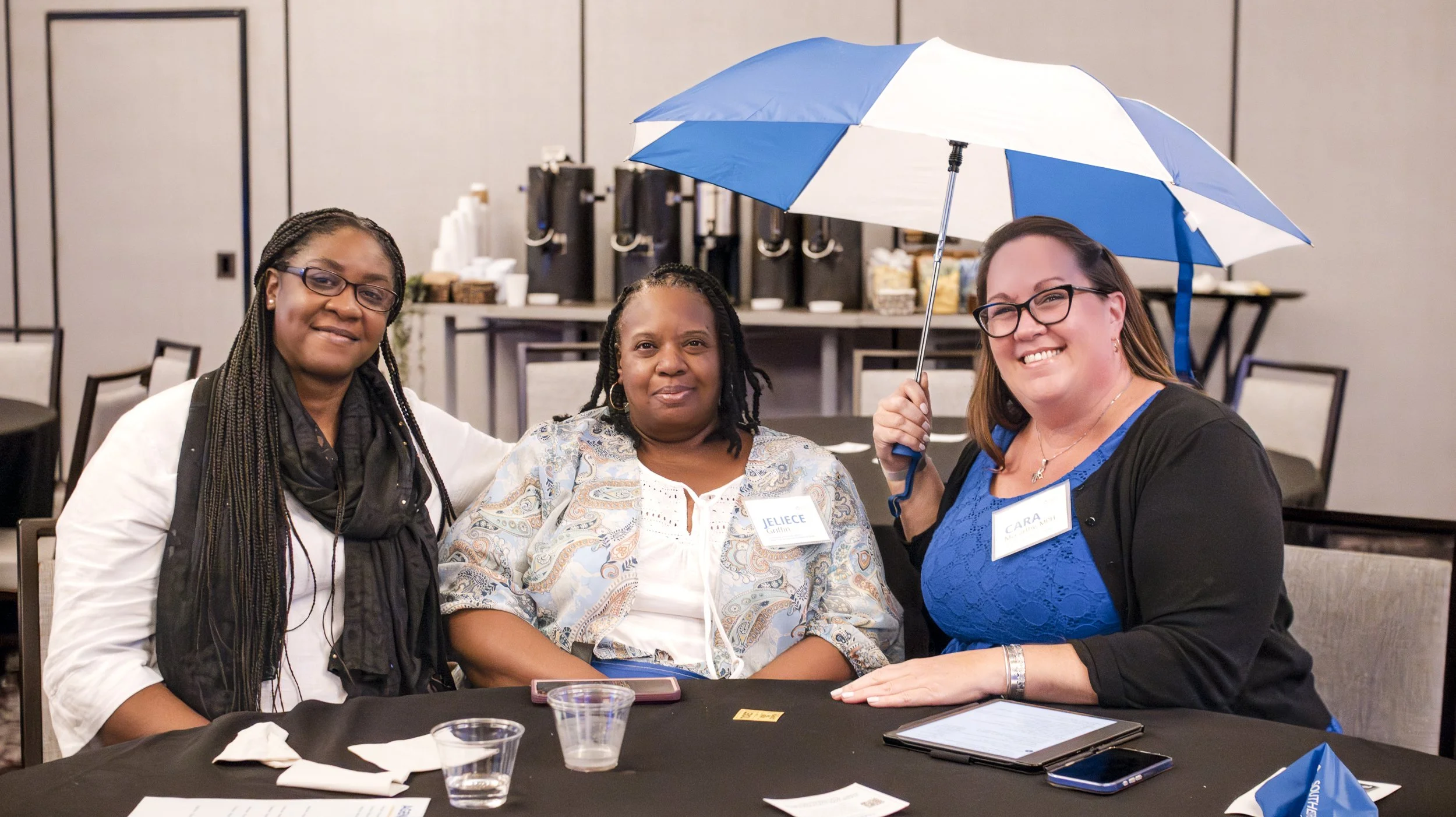 Three women sitting at a table during an event, one woman on the right holding a blue and white umbrella, all smiling, with name tags that read Jelice and Cara.