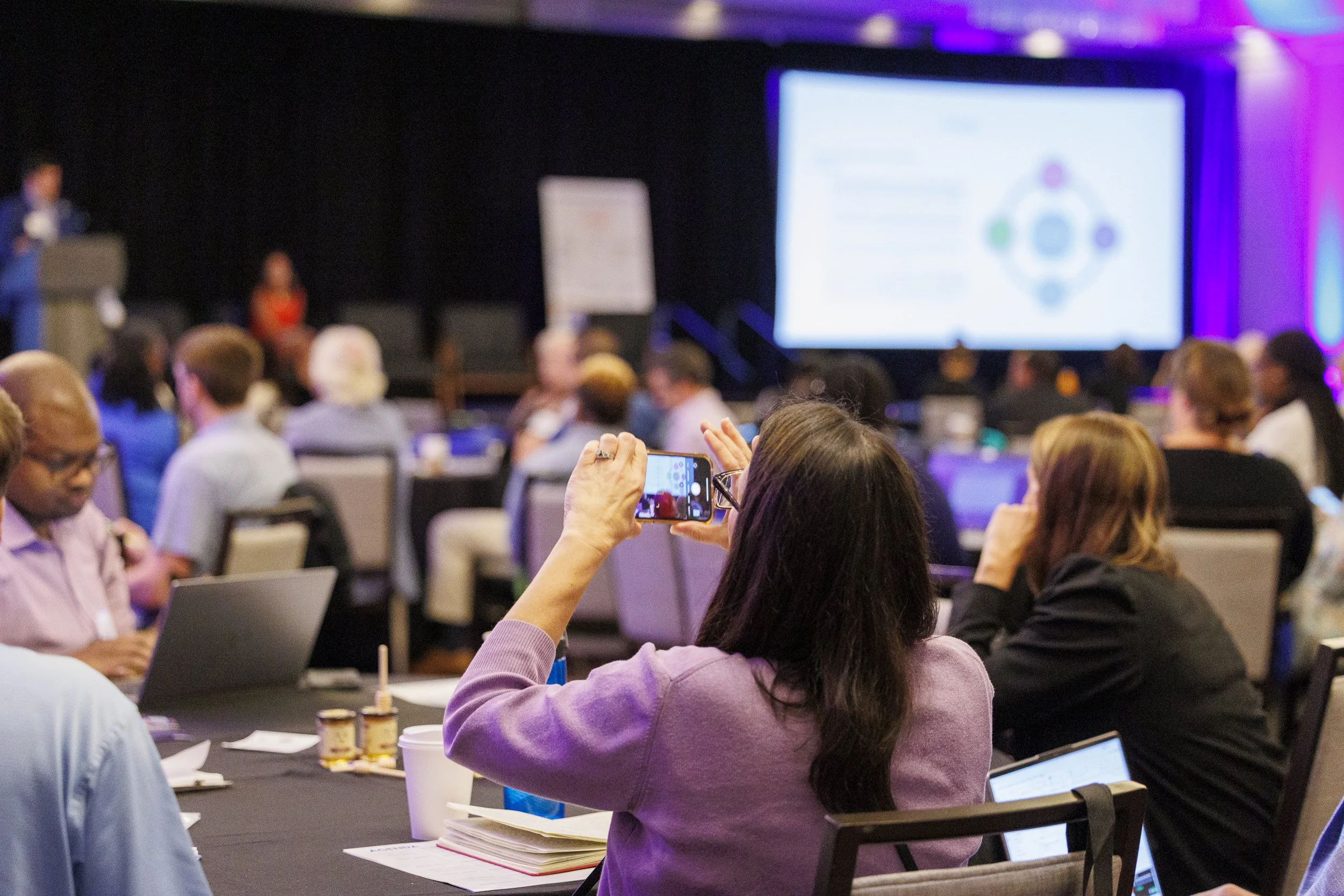 A woman taking a photo with her phone during a conference or seminar, with a large presentation screen in the background. Other attendees are seated at tables, some using laptops.