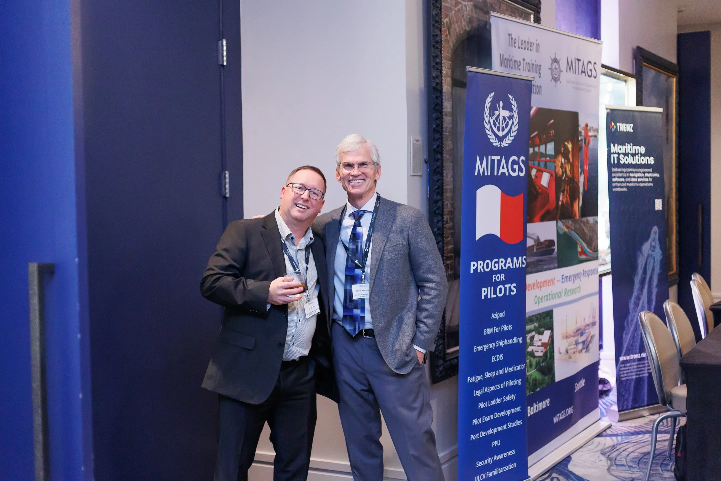 Two men in suits smiling and posing together at a maritime training conference, with banners in the background advertising programs for pilots and maritime IT solutions.