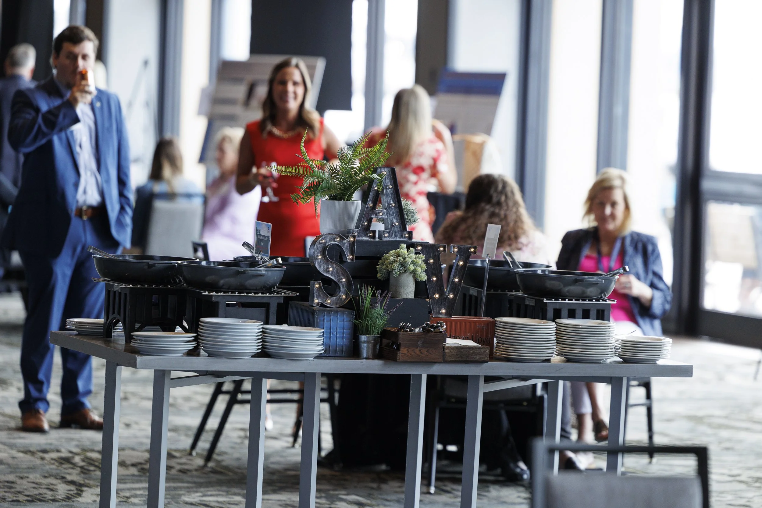 Buffet table with plates and decorative items at a social gathering in a modern venue with large windows and people in business attire in the background.