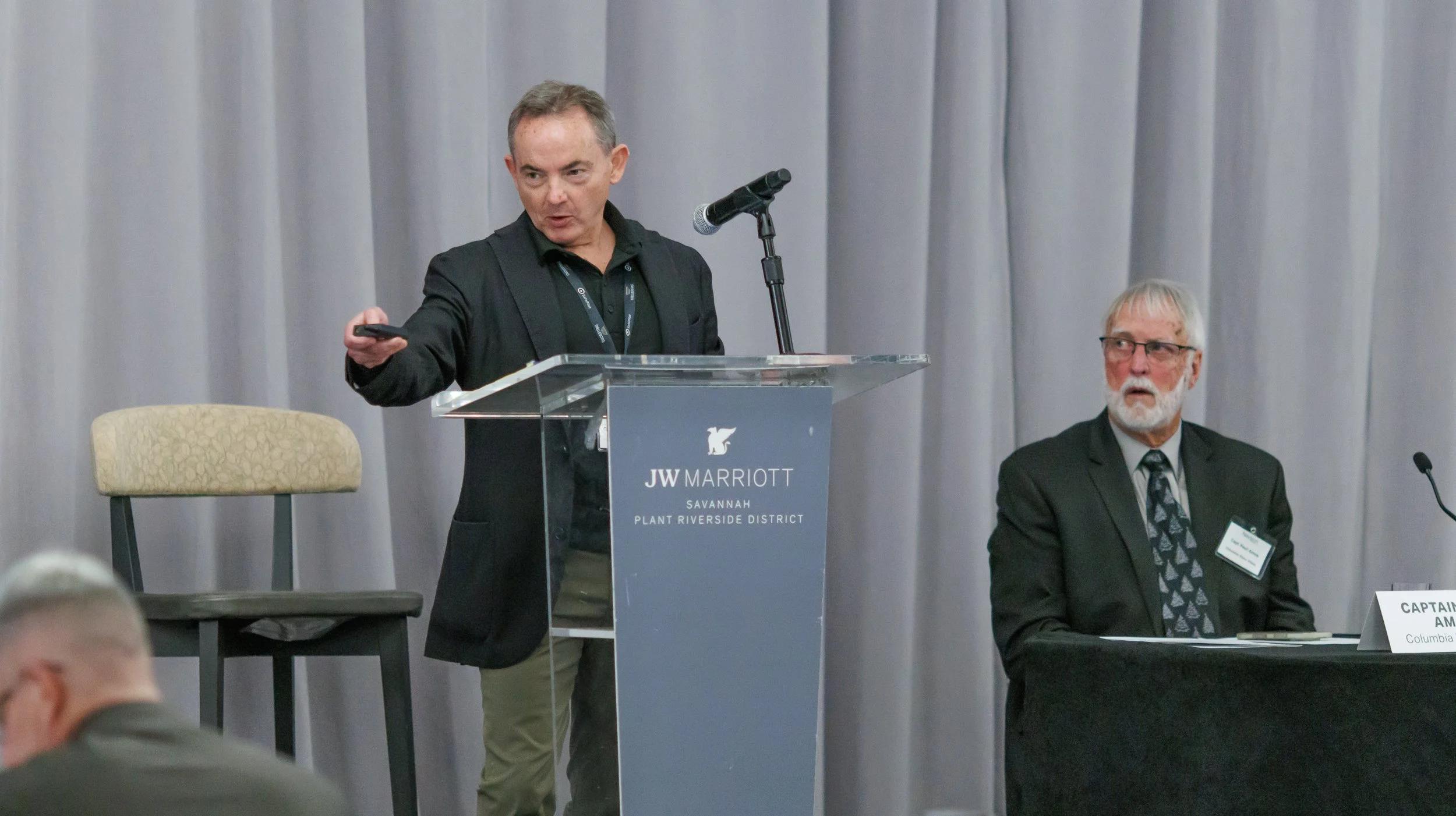 A man in a black blazer and black shirt speaking at a podium with a microphone at a conference, with a seated man with white hair and a beard listening nearby, in a room with gray curtains and a beige chair.