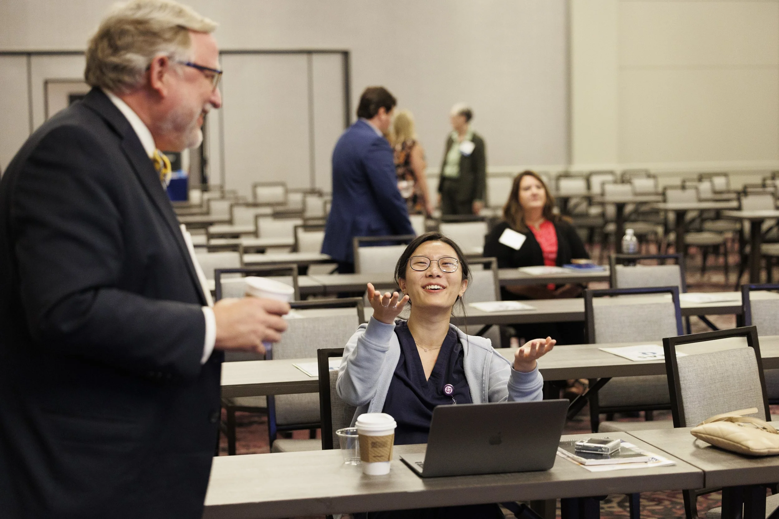 A young woman with glasses sitting at a conference table, smiling and gesturing with her hands, while a man in a suit stands nearby holding a coffee cup. Other people are in the background in a conference room setting.