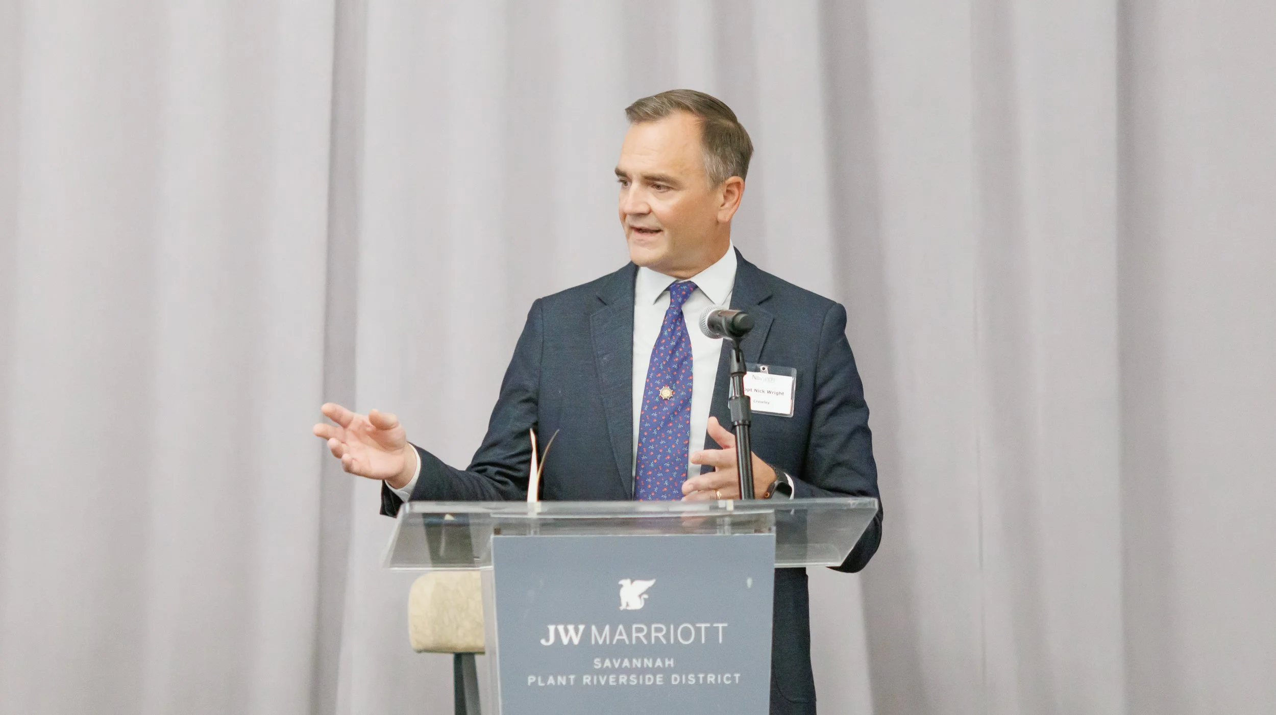 A man in a dark suit and tie speaking at a podium with a JW Marriott sign, in front of a gray curtain.
