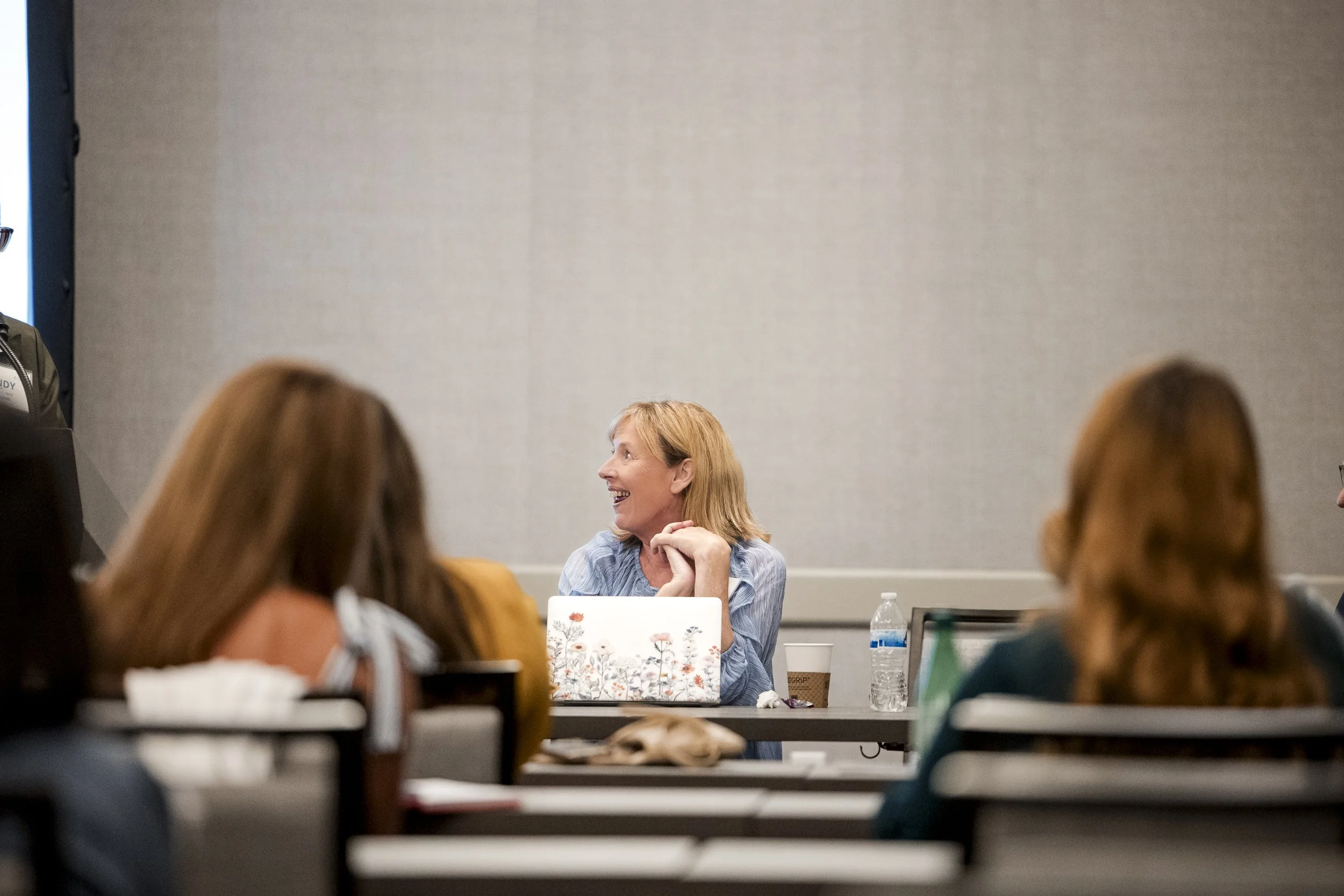 A woman with blonde hair smiling and looking to her left at a conference or meeting, with people seated in front of her and a laptop on the table.