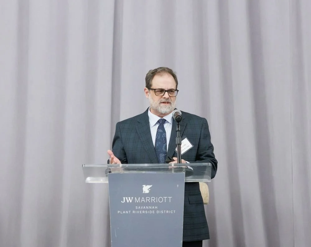 A man in a suit and tie is speaking at a podium with a microphone. The podium has a sign that reads JW Marriott Savannah Plant Riverside District.