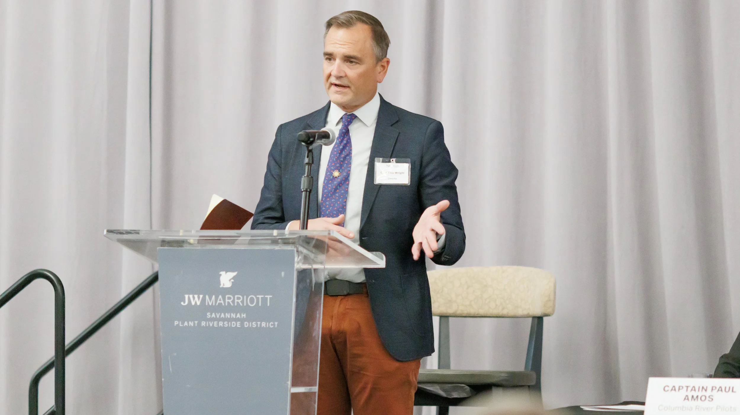 A man in a dark suit jacket, white shirt, and patterned tie stands behind a clear podium with the JW Marriott logo, speaking to an audience with a microphone in front of a gray curtain backdrop. A name tag is visible on his jacket.