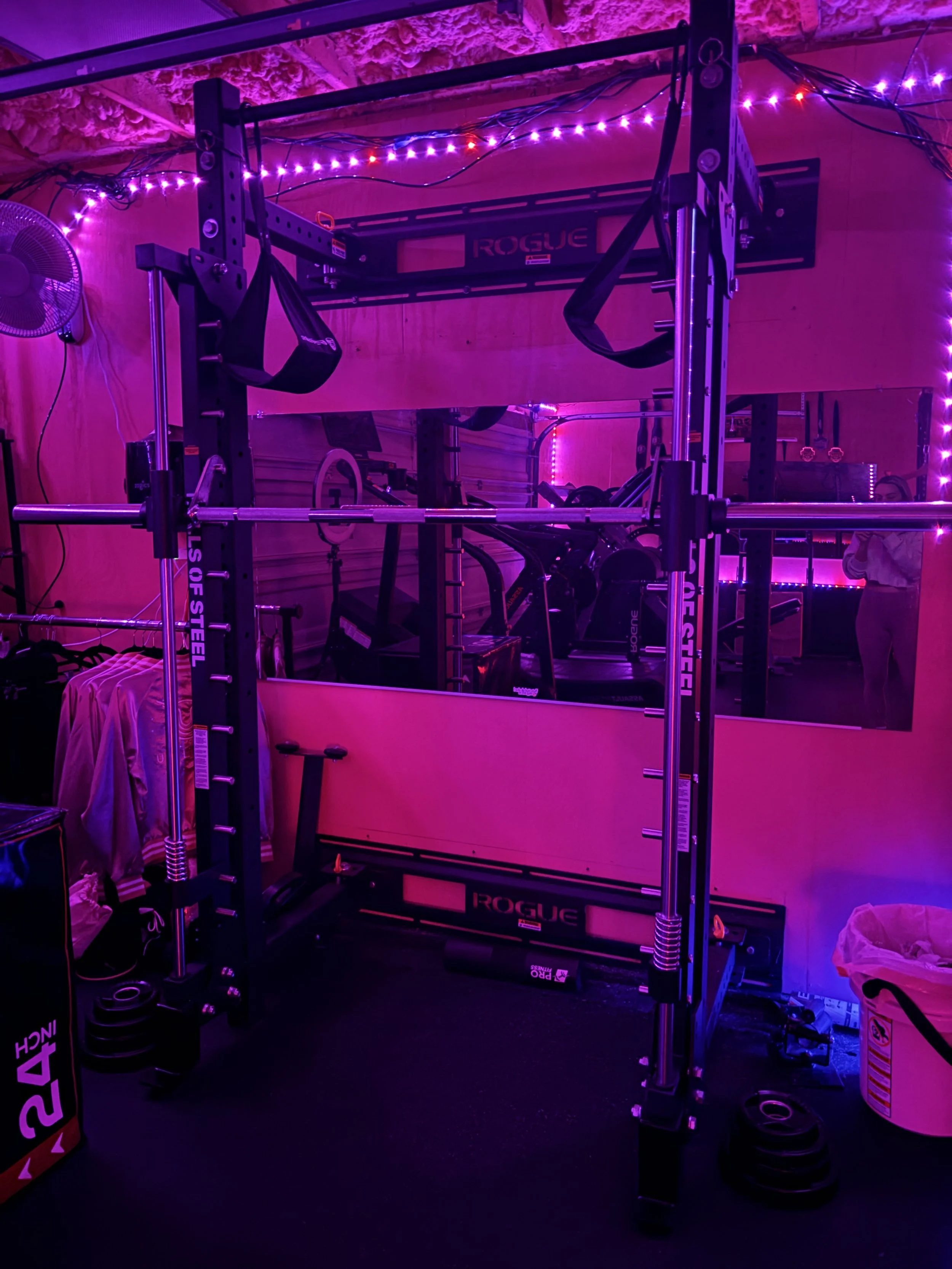 Gym with black Rogue fitness power rack illuminated by purple and pink LED strip lights, with barbells, weights, and an exercise ball nearby, and a person taking a mirror selfie.