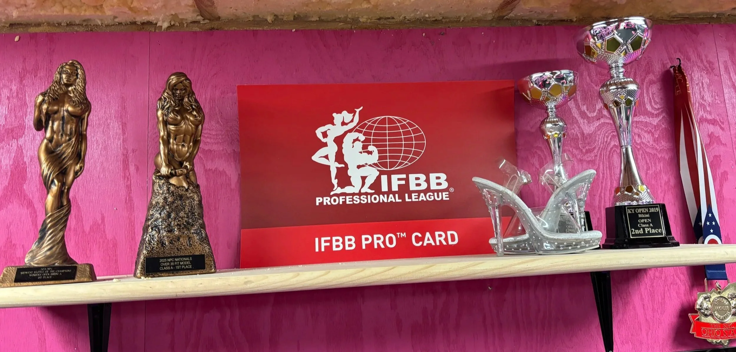 Awards shelf with trophies, a promotional card, and medals against a pink wall. Trophies include two bronze statues and silver cups. The card displays the IFBB logo and text for professional league achievements.