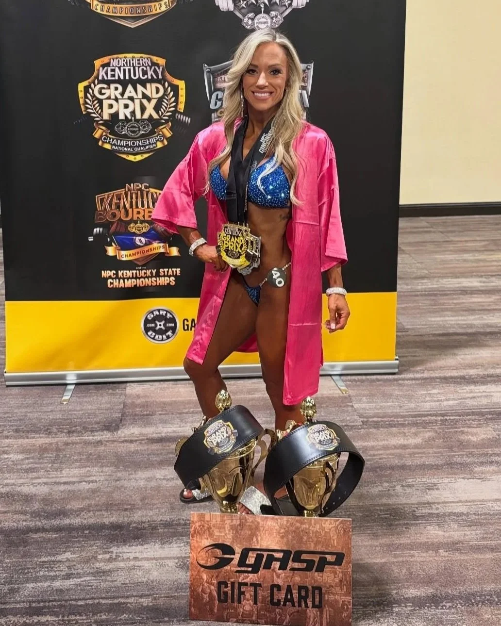 A woman at a bodybuilding competition shows her trophies and medals. She is wearing a blue bikini with a pink robe, gold trophies on her feet, and medals around her neck. Behind her is a black and yellow banner with competition logos and text.