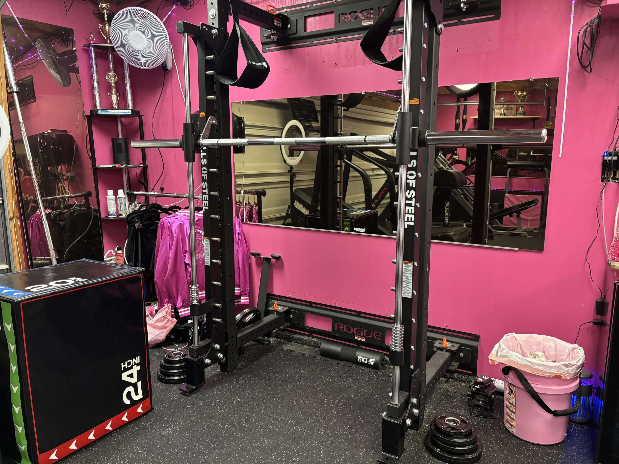 A fitness gym with a black squat rack, pink walls, and various workout equipment. There is a large mirror on the pink wall, and a pink trash can on the floor. A fan, some clothing, and water bottles are visible, along with a black plyometric box and 