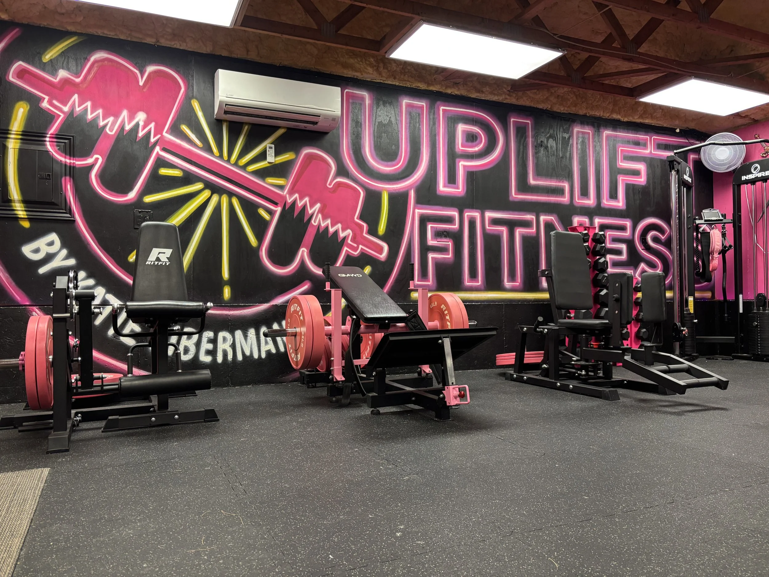 A gym with a colorful neon sign on the wall reading "UPLIFT FITNESS" and a pink ice cream cone graphic. The gym has various workout equipment including weightlifting machines and benches, and the floor is carpeted with black rubber mats.