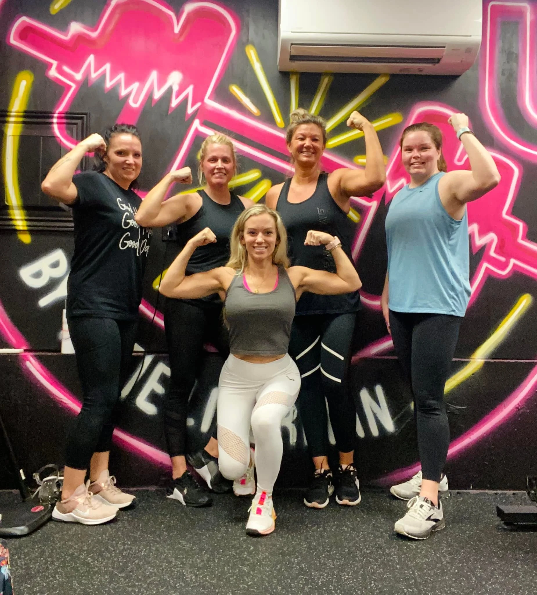 Group of five women flexing their arm muscles in a fitness studio with a black wall decorated with neon pink and yellow graphics in the background.