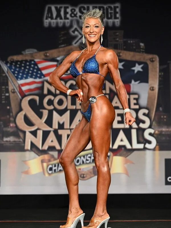 A female bodybuilder on stage at a national bodybuilding competition, posing in a blue bikini with high heels, with a backdrop featuring the American flag and event branding.