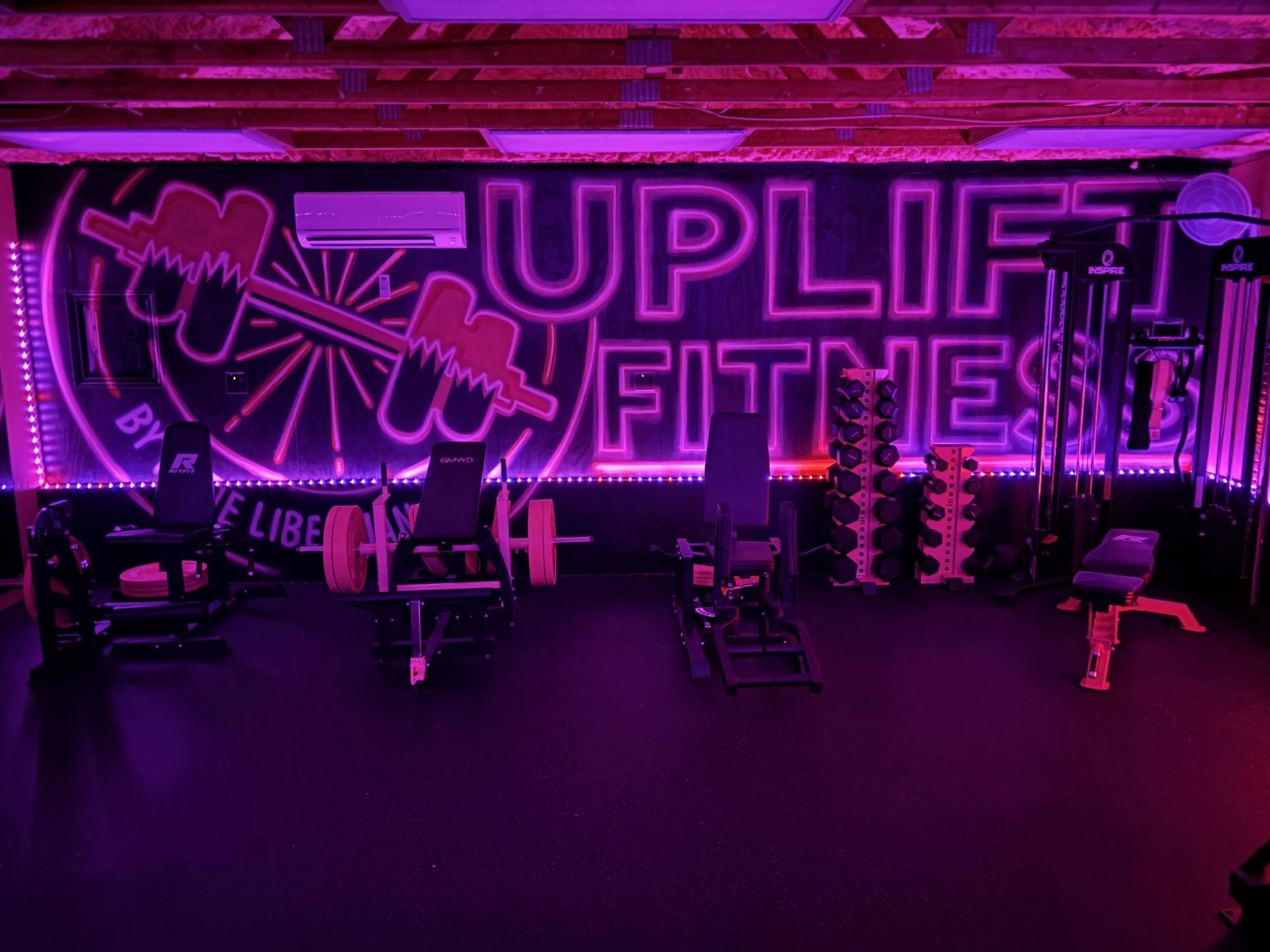 A gym with neon purple and pink lighting, featuring workout equipment including weight benches, dumbbells, and a treadmill. The wall has a large neon sign that reads 'UPLIFT BY FITNESS' with an illustration of a weightlifting bar.