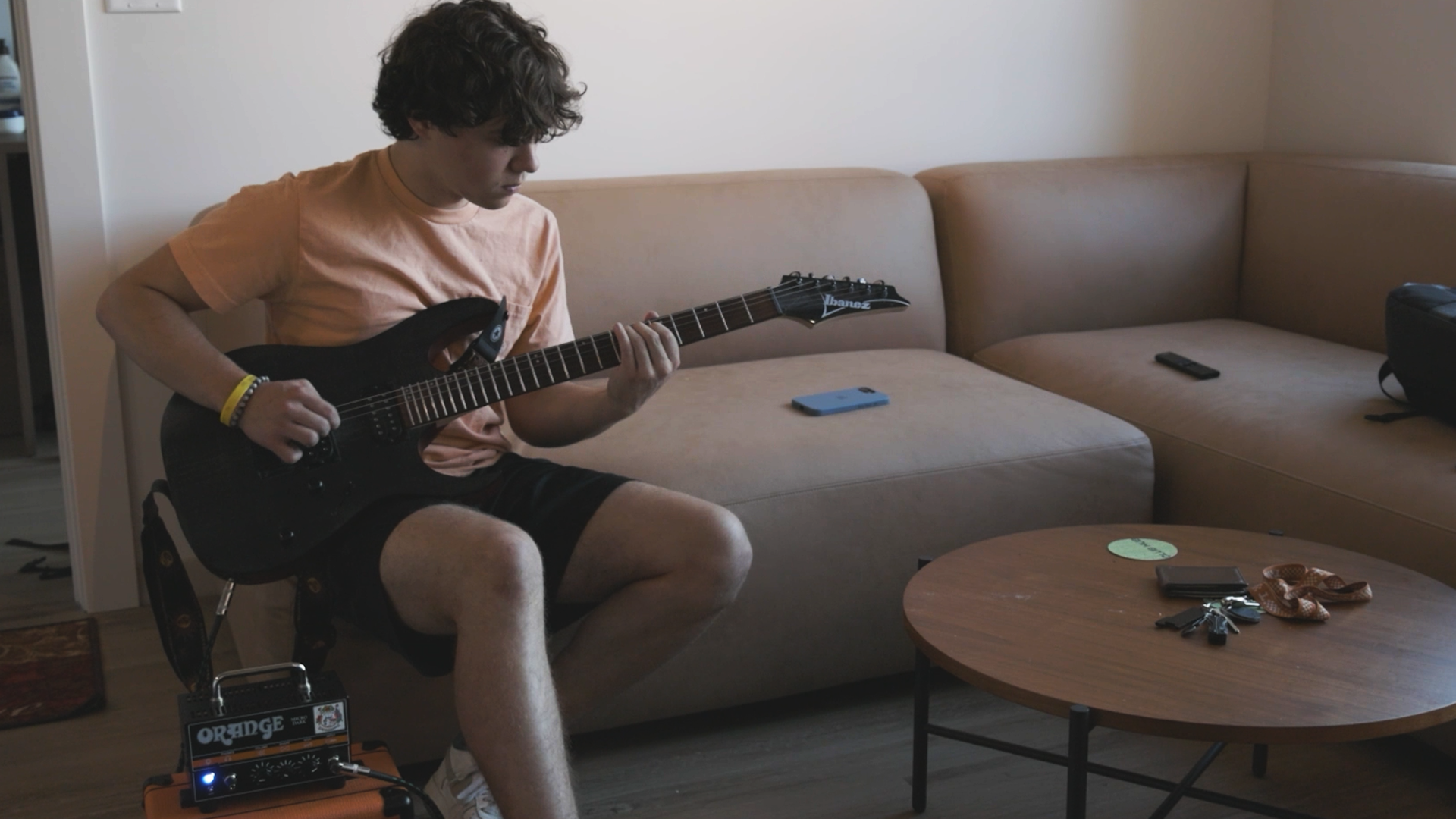 A young man with curly hair wearing a peach T-shirt and black shorts sitting on a beige sofa, playing a black electric guitar connected to an orange Orange brand amplifier, in a living room with a wooden coffee table, a blue smartphone, a black pouch, keys, and a green sticker on the table.