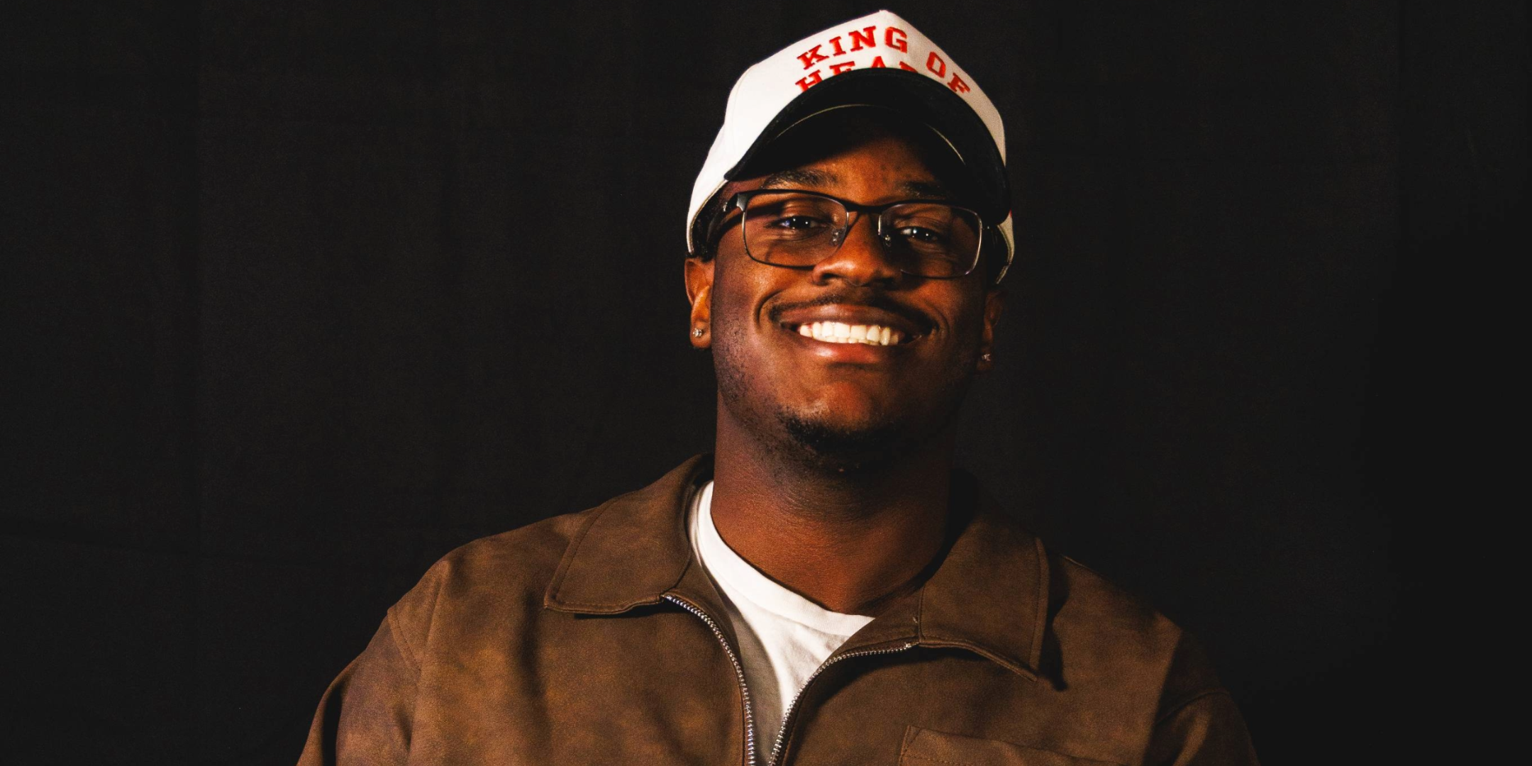 A young man smiling with glasses, wearing a white hat with red text and a brown jacket, against a black background.
