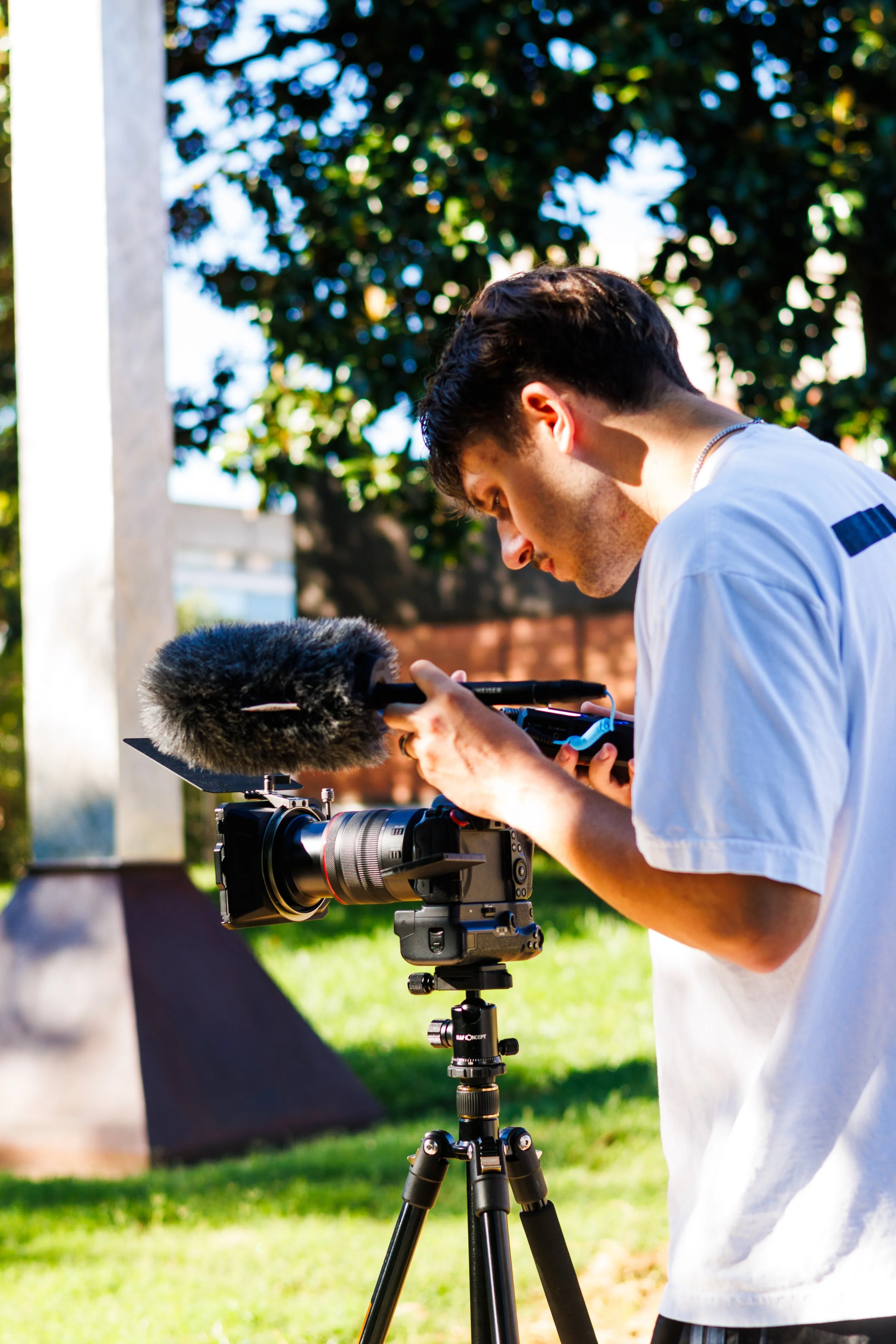 A young man using professional camera equipment with a microphone attached, outdoors in sunlight with a tree and grass background.
