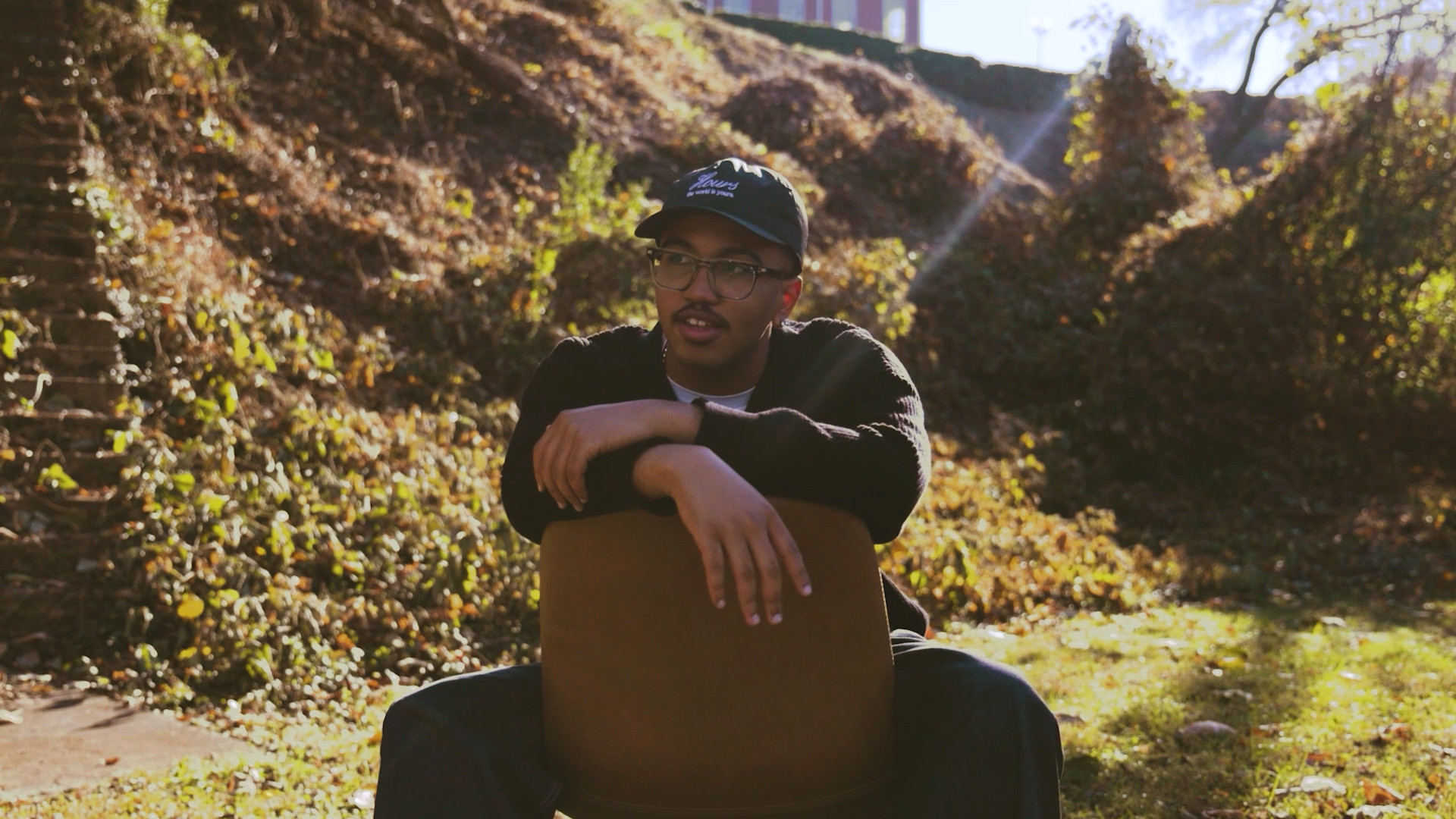 A young man with glasses and a cap sits outdoors leaning on a chair backrest in a natural setting with sunlight and bushes in the background.