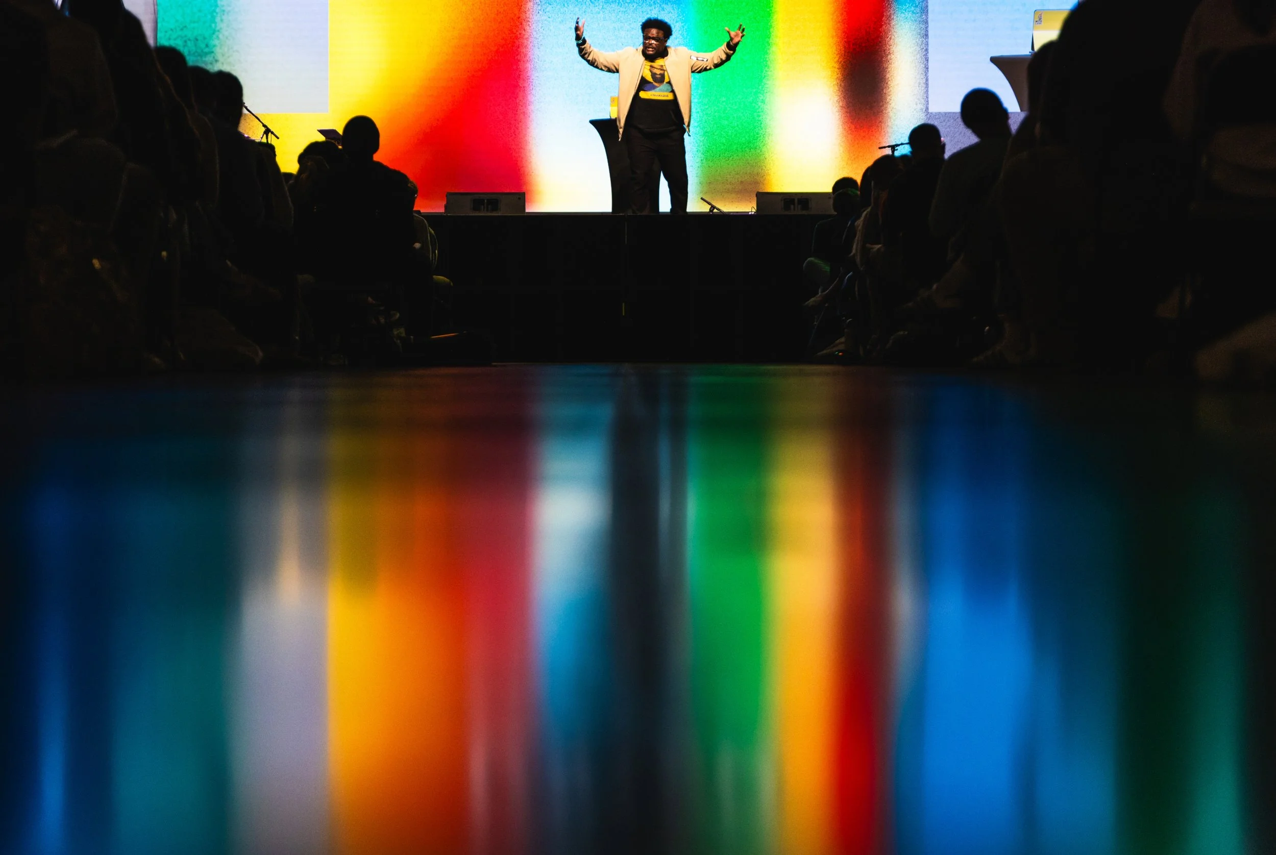 A person standing on stage with arms raised, speaking to an audience, in front of a colorful background, with silhouettes of audience members visible.
