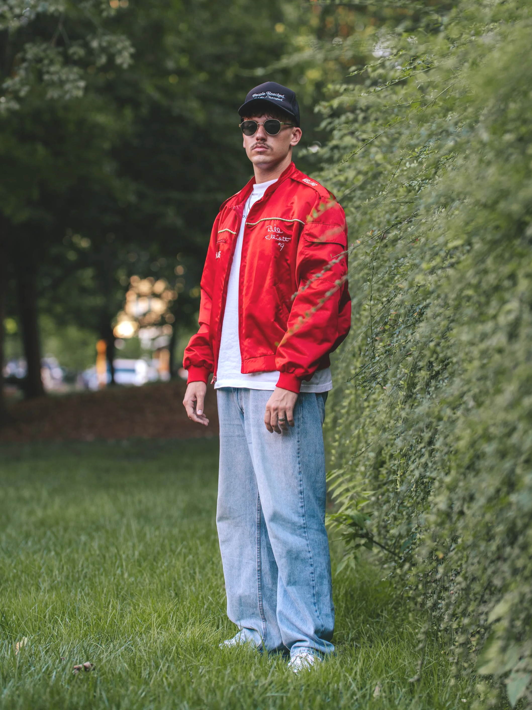 A young man wearing sunglasses, a black cap, a red satin bomber jacket, a white t-shirt, light blue jeans, and white sneakers, standing outdoors beside a green hedge with trees in the background.