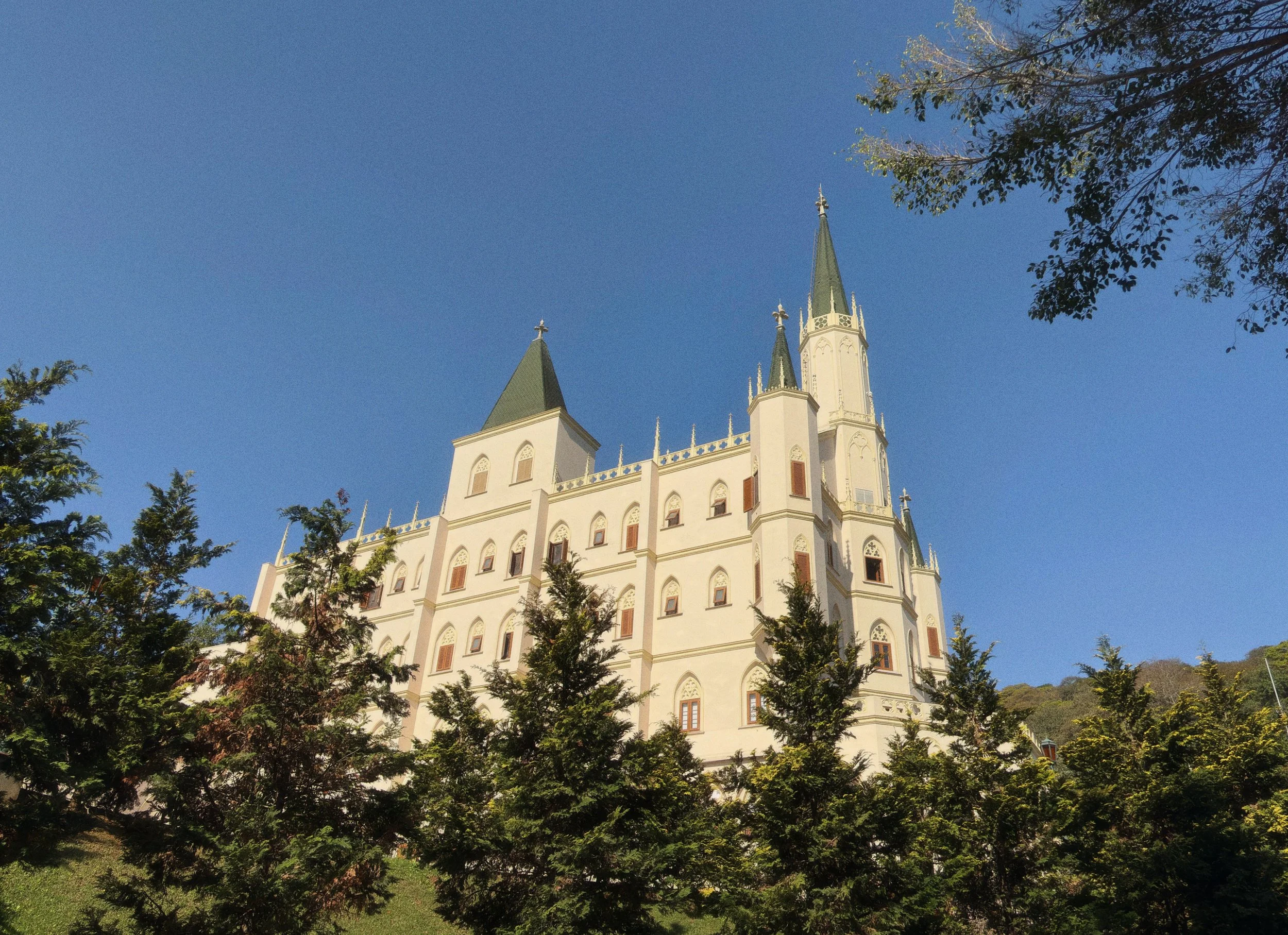 Castelo branco com torres altas e janelas decorativas, sob céu claro com árvores ao redor.