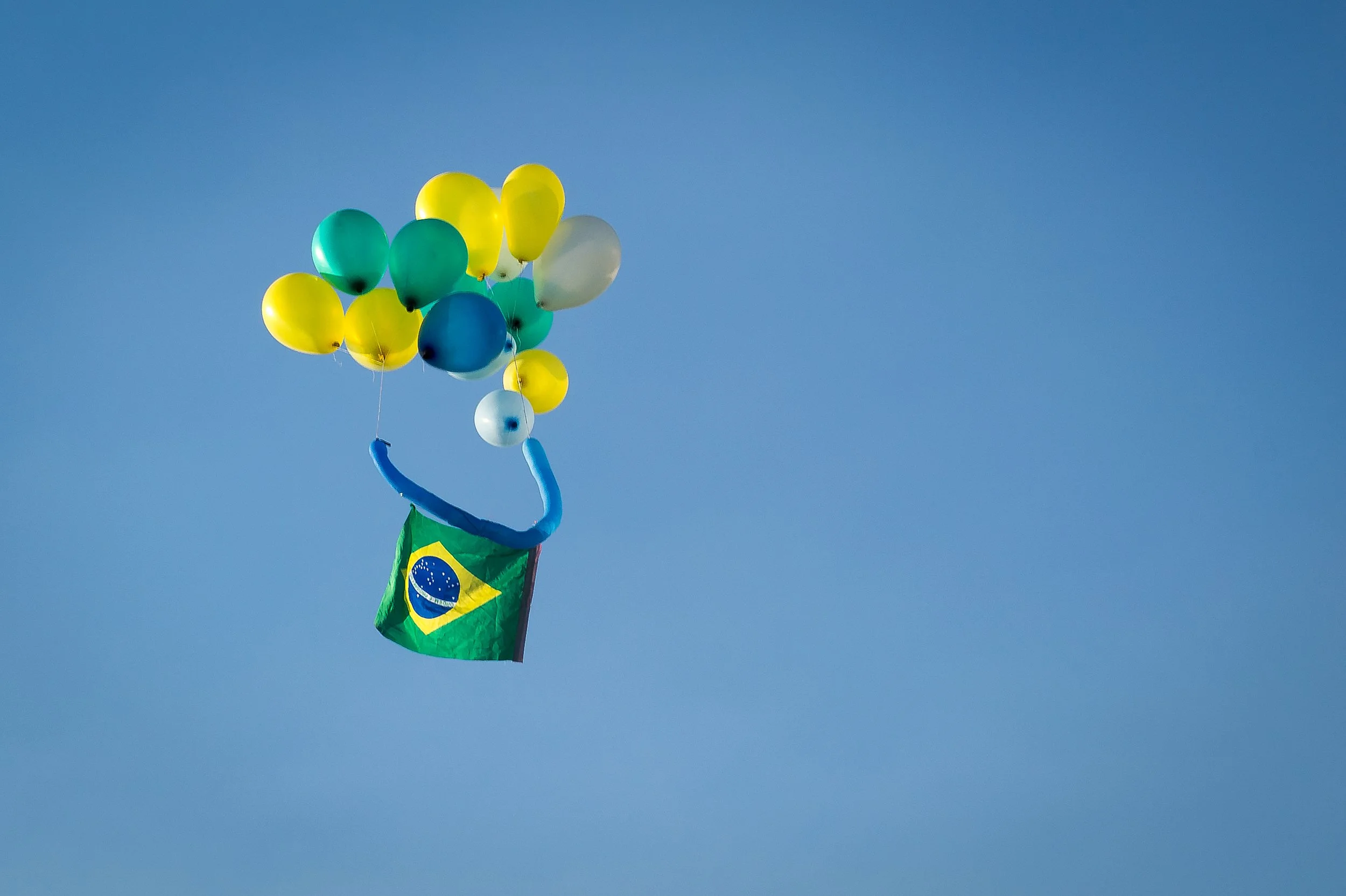 Balões coloridos formando um sorriso com uma bandeira do Brasil pendurada, flutuando no céu azul.