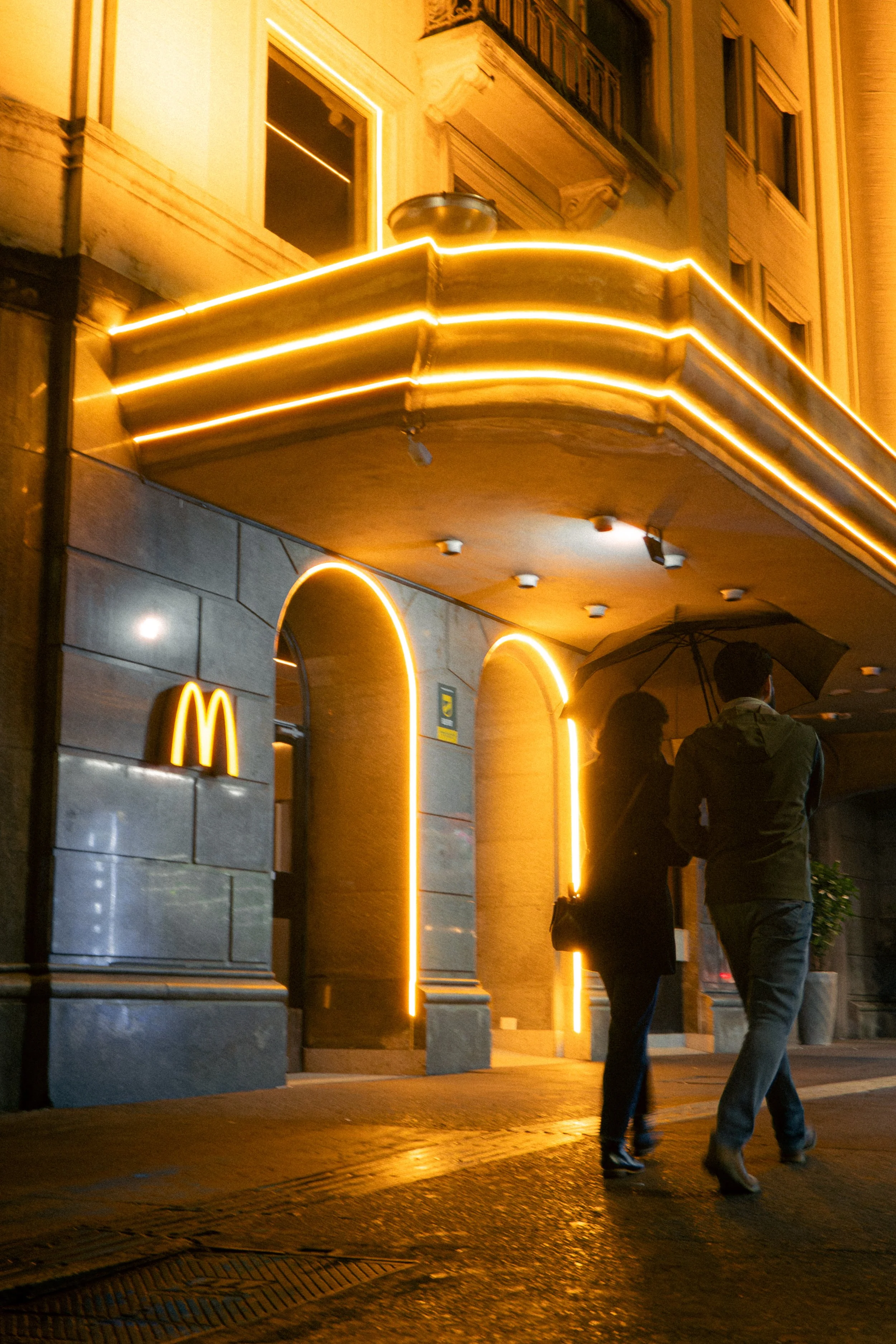 Caminhada de duas pessoas sob guarda-chuvas em frente a uma entrada de restaurante ou loja iluminada com faixas de luz amarela ao longo de um edifício.