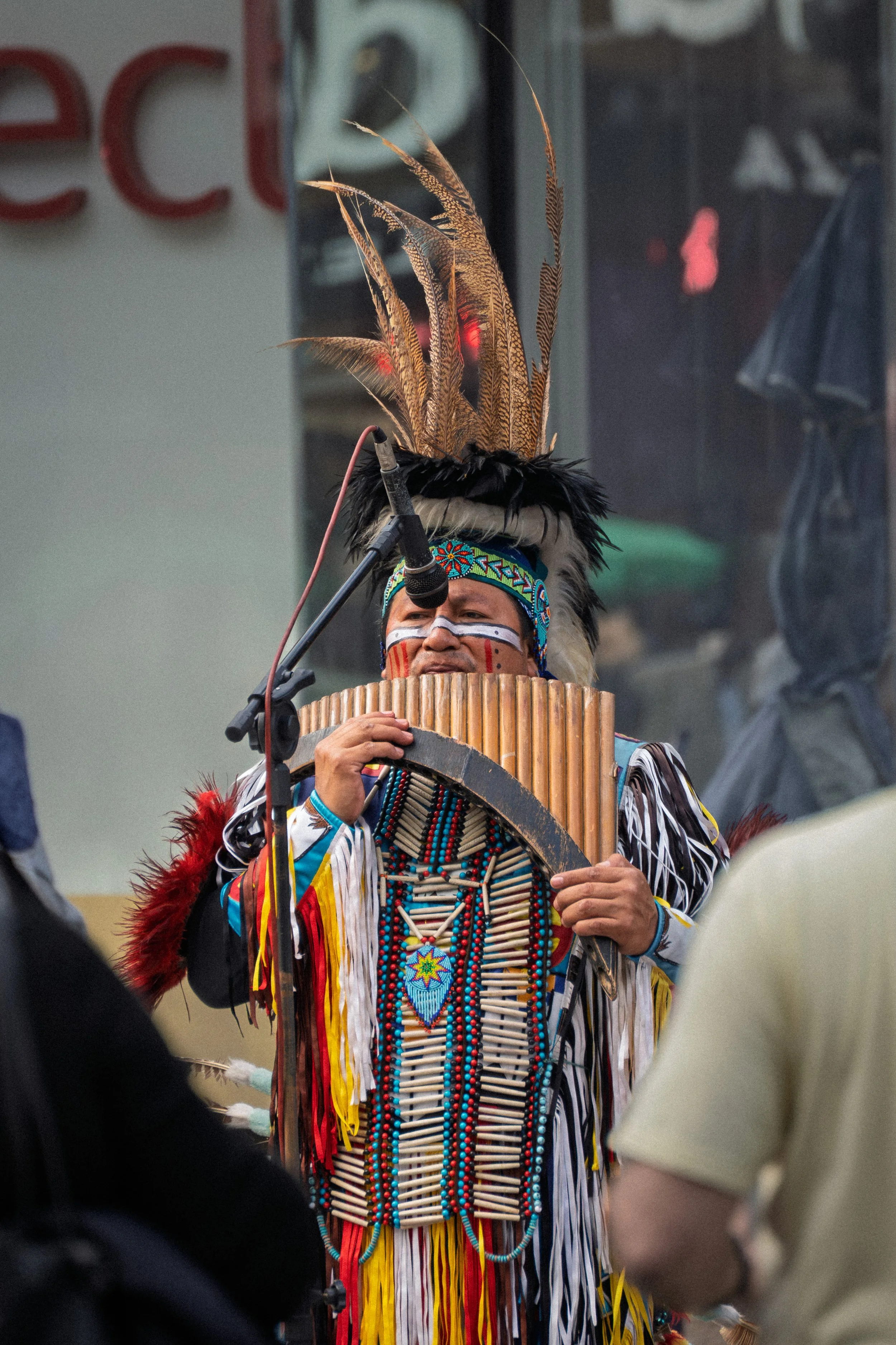 Homem indígena vestindo trajes tradicionais com penas e colares, tocando um instrumento musical, durante uma apresentação ou celebração.