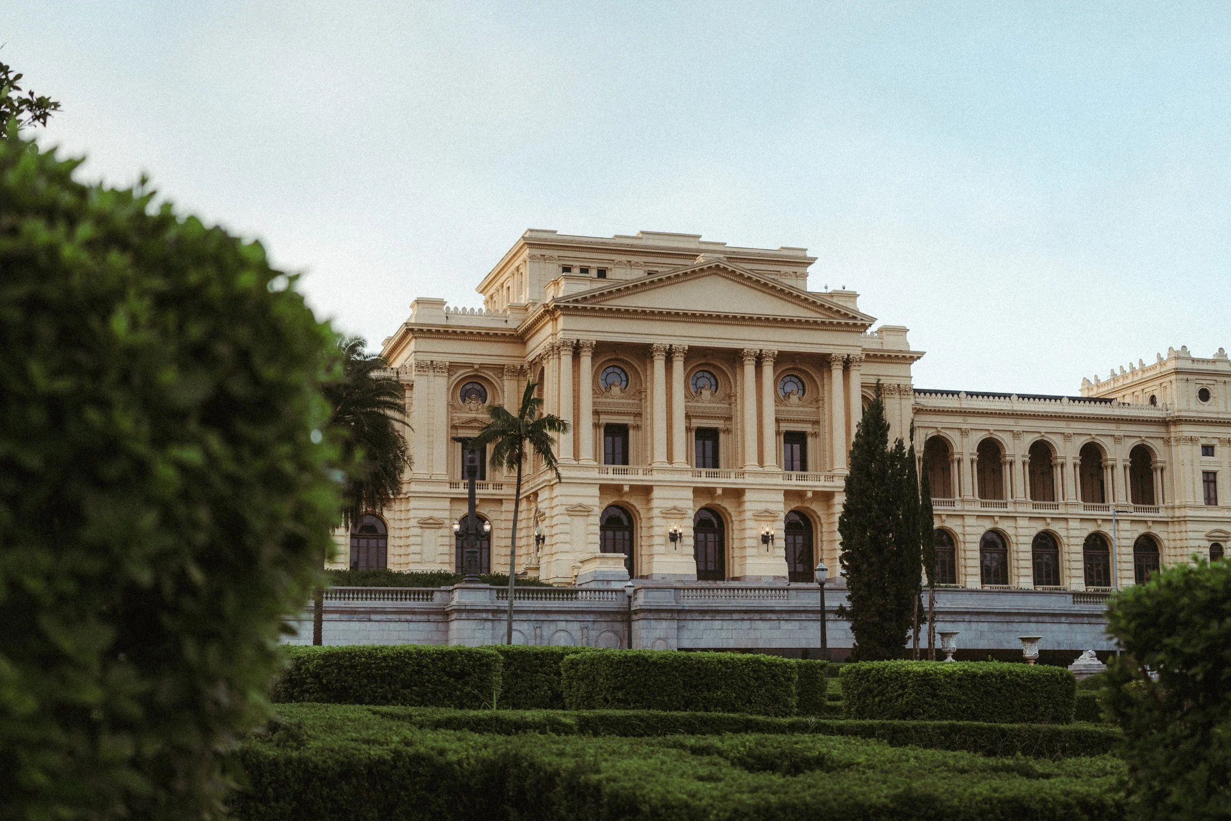 Prédio grande de arquitetura clássica, com colunas e detalhes ornamentais, rodeado de jardins bem cuidados com arbustos e árvores, ao entardecer.