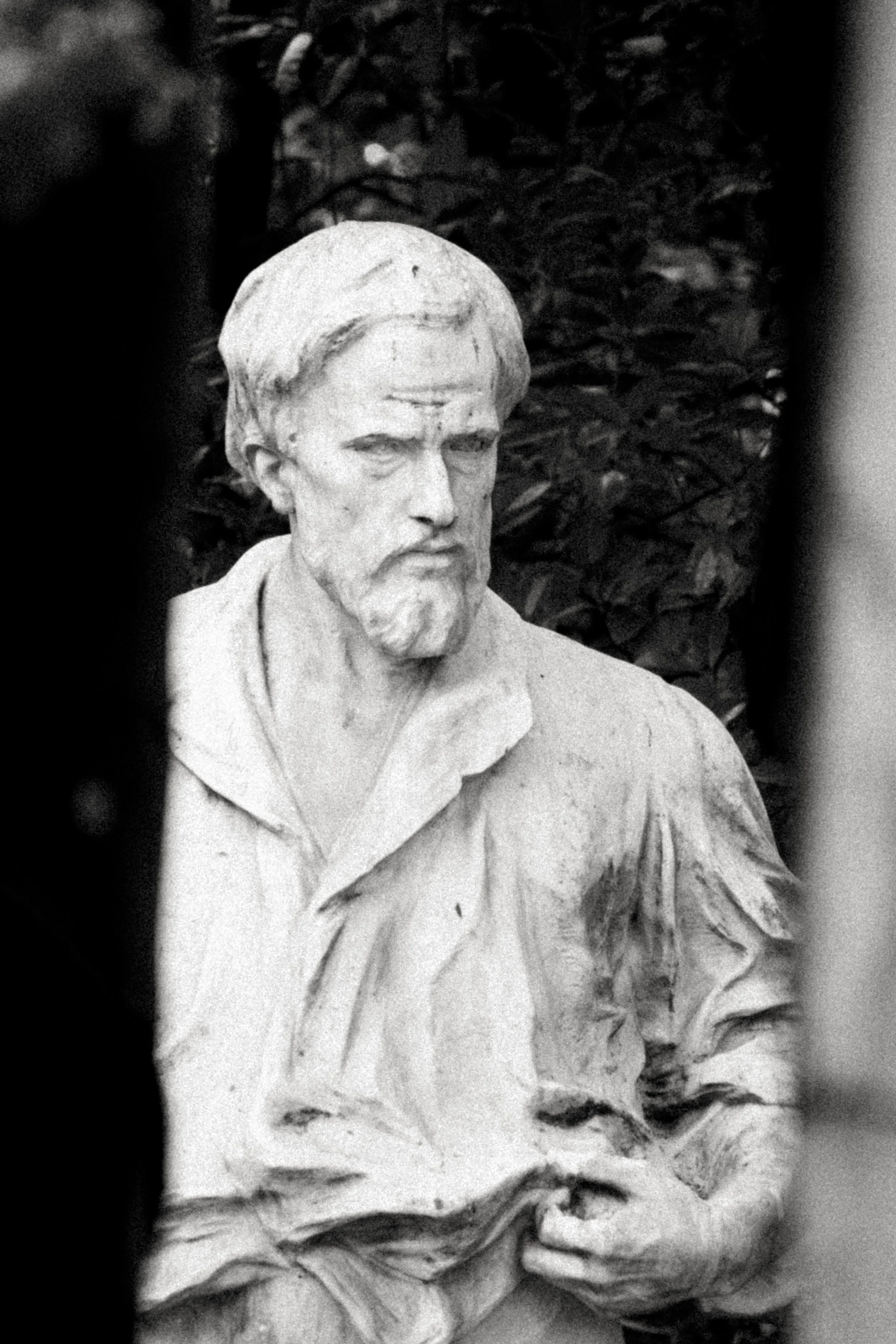 Escultura de uma figura masculina com barba e cabelo curto, usando uma camisa com gola, em um fundo de vegetação.