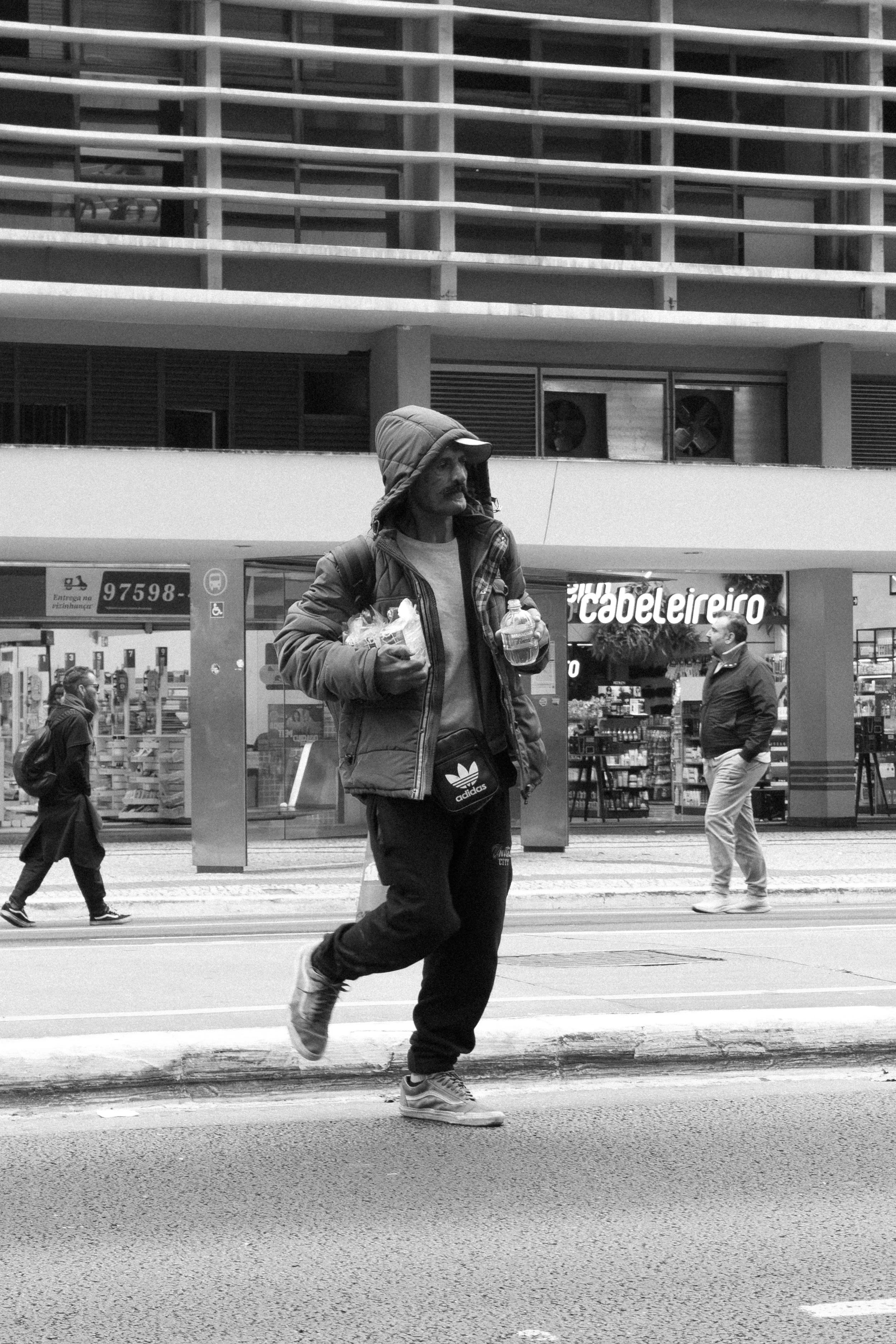 Homem andando na rua carregando duas garrafas de água, usando jaqueta e boné, com outras pessoas ao fundo na frente de uma loja de materiais de construção.