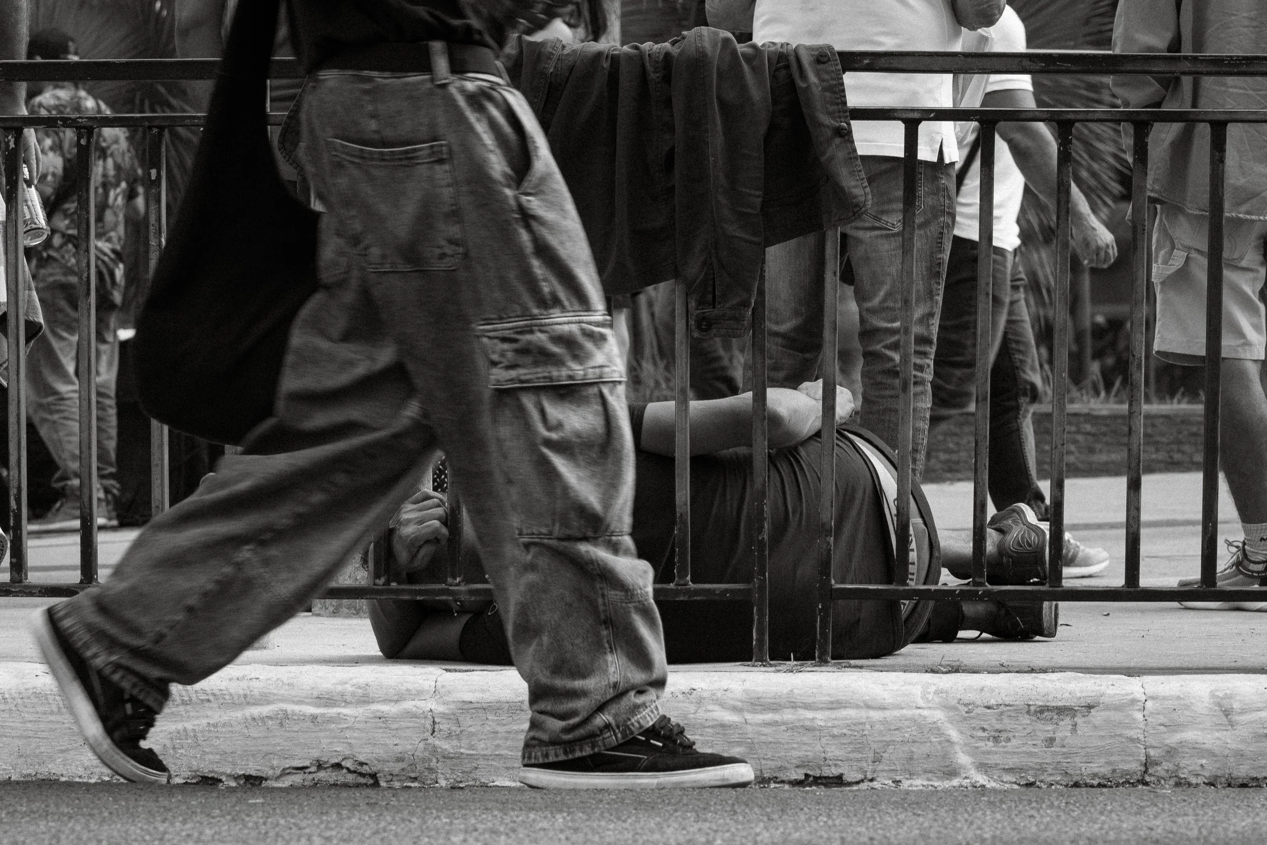 Pessoa caída ao lado de uma grade na rua, cercada por outros pedestres, em uma cena em preto e branco.