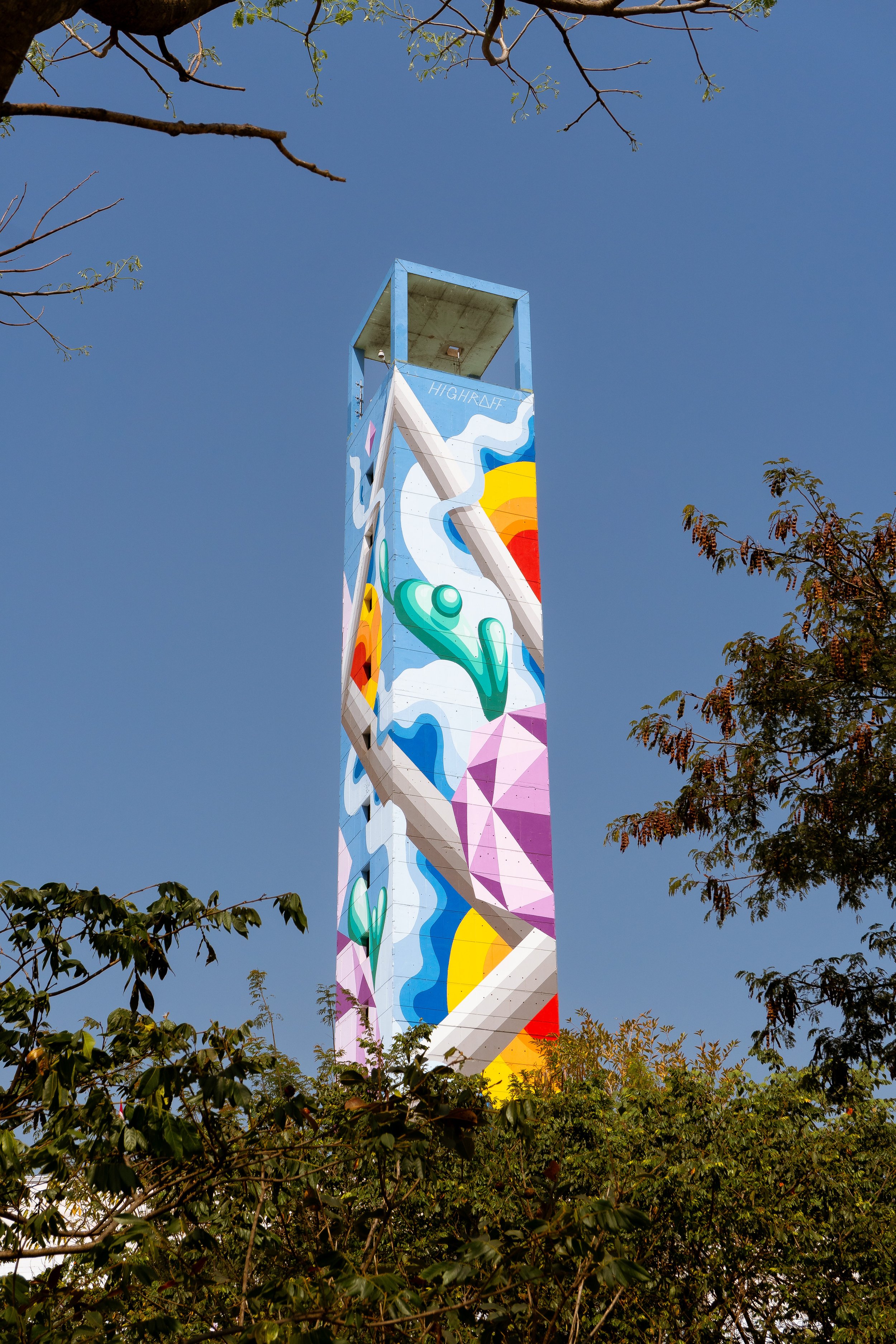 Torre de escalada decorada com arte colorida e formas geométricas, contra céu azul, com árvores ao redor.