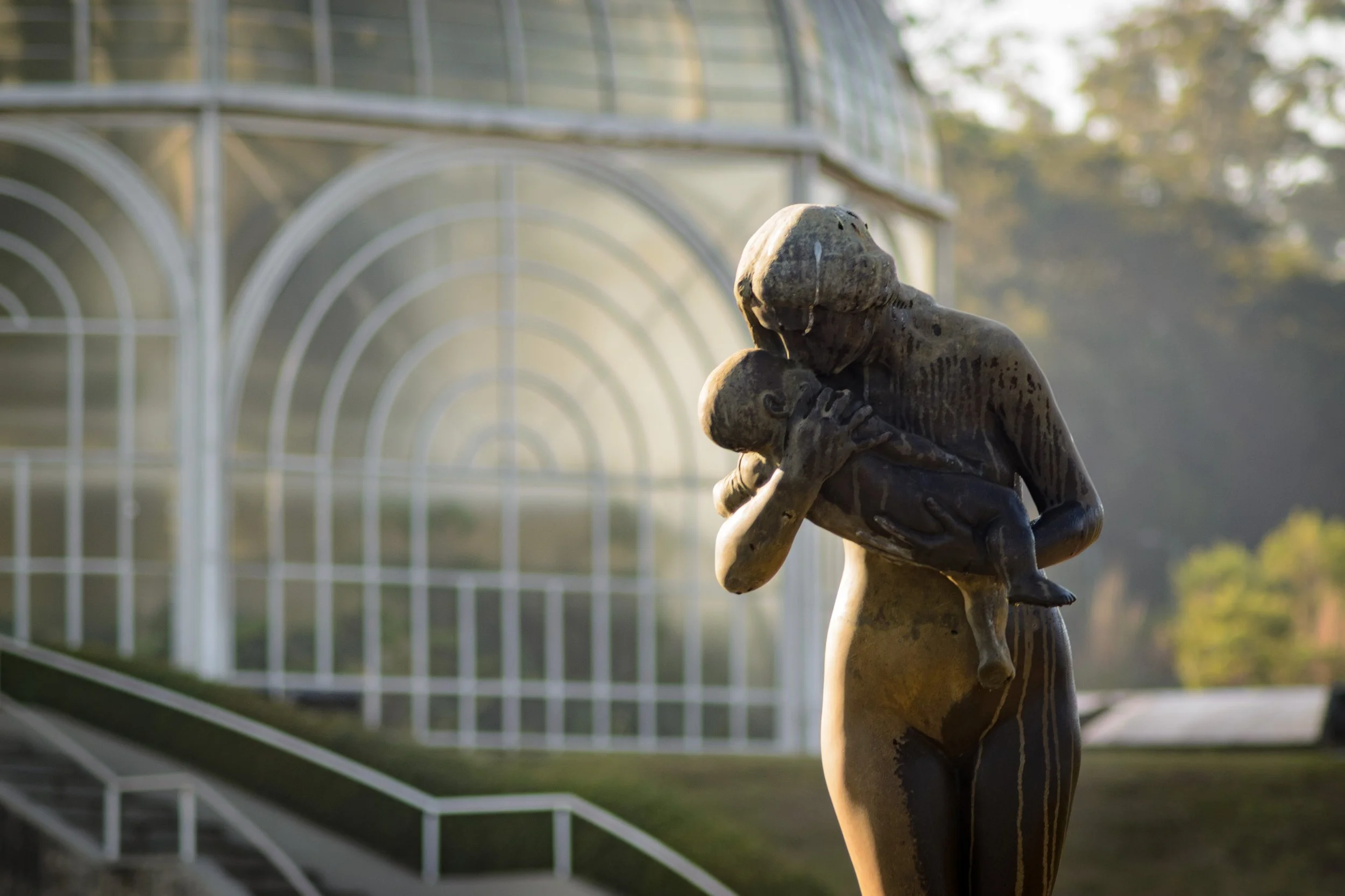 Escultura de uma mulher segurando uma criança, ao ar livre com estrutura de vidro ao fundo.